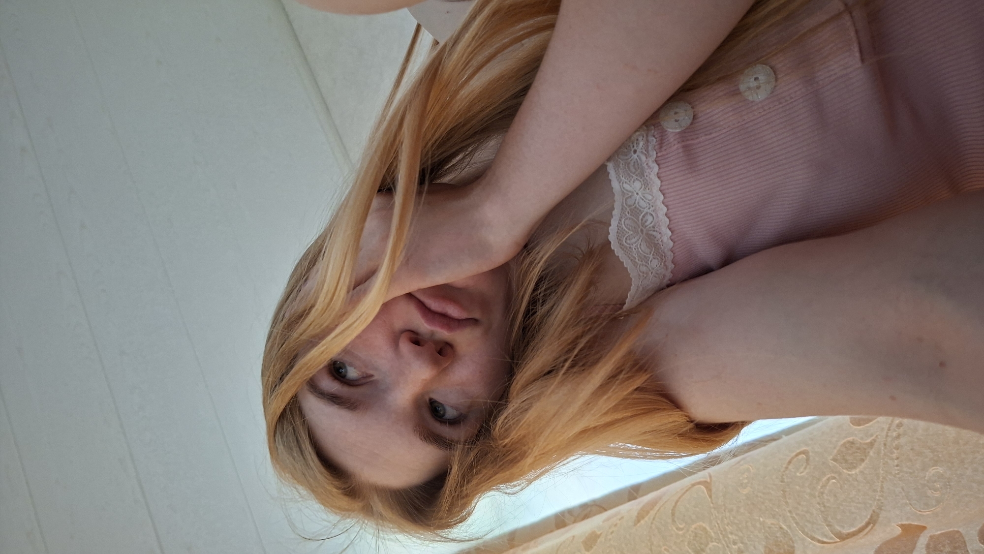 This image presents a candid, low-angle portrait of a young woman. Due to the photo's rotation, her head is positioned towards the left side of the frame, and her body extends towards the right. She has long, straight blonde or light brown hair that cascades around her face. She is wearing a light pink top featuring white lace trim along the neckline and two decorative white buttons on the placket. Her left hand is raised, partially obscuring the upper part of her face, with her eyes gazing directly upwards towards the camera. Her lips are slightly parted, and her expression conveys a sense of shyness or contemplation.
The setting appears to be indoors. The background behind her upper body and head shows a light-colored, possibly paneled wall or ceiling. Towards the bottom right of the frame, a segment of a patterned, light-toned fabric, likely a curtain or blanket, is visible. The soft, natural lighting suggests the photo was taken during the daytime. There is no visible text in the image. This photograph was taken in Большой Букор, Russia.