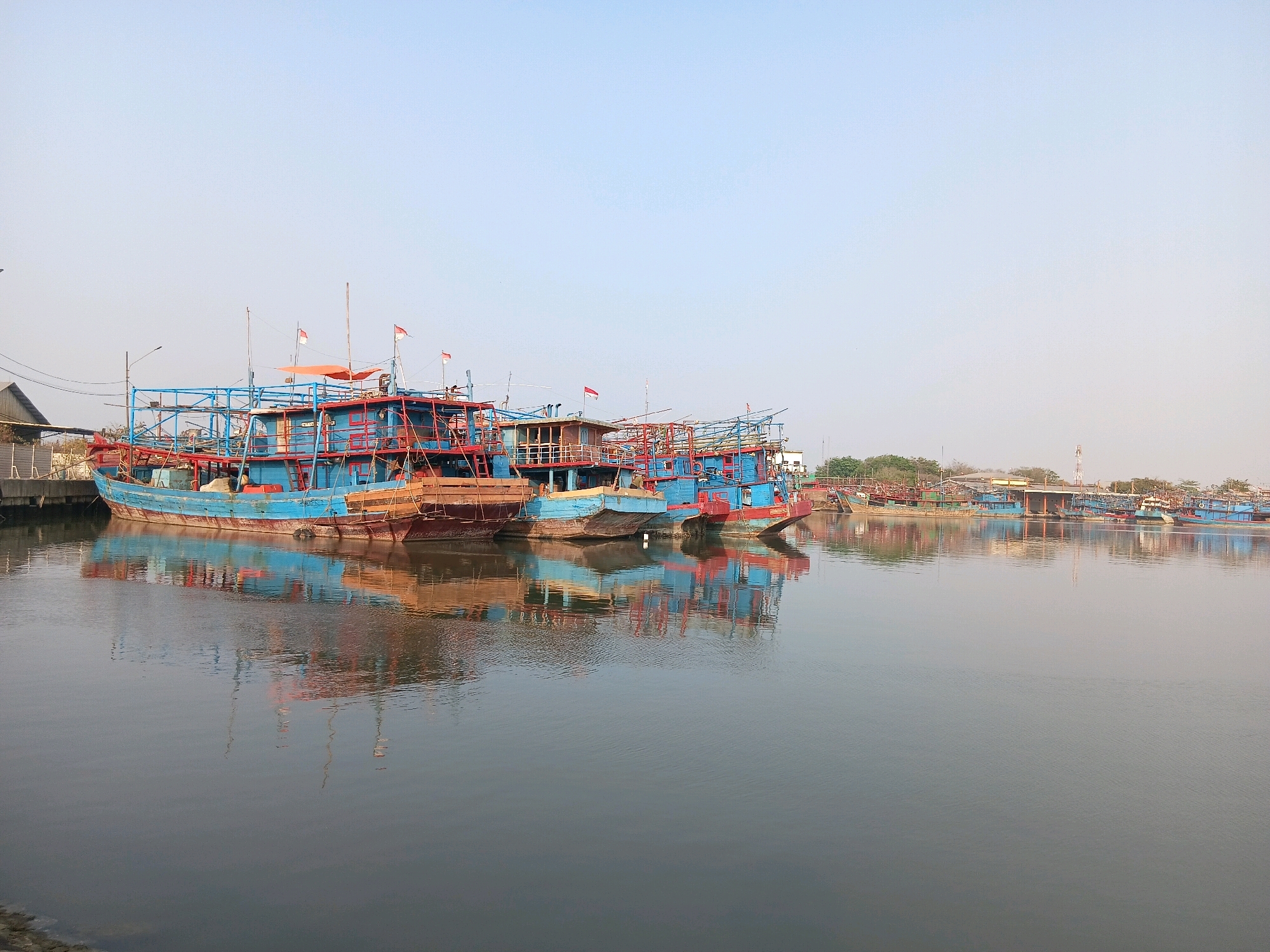 The image depicts a harbor in Cirebon, Indonesia, filled with numerous blue and red fishing boats docked along a quay. The boats, varying in size and construction, are equipped with multiple decks and rigging, some flying small red and white flags. The water in the foreground is calm, reflecting the boats and the pale blue sky. In the background, a low-lying cityscape with buildings, trees, and some industrial structures is visible, suggesting an urban coastal environment. The overall scene is one of a working harbor, with boats at rest.