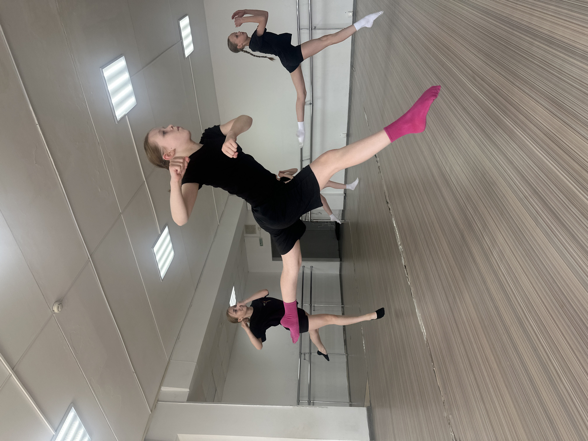 The image depicts a dance studio in Votkinsk, Russia, with three young female dancers in mid-motion. They are wearing black tops and shorts. One dancer has pink socks, while another has black socks and the third has white socks. The studio features a barre along the wall and a mirrored wall, reflecting the dancers and the room. The floor is made of light brown wood planks. The ceiling is white and has fluorescent lights.