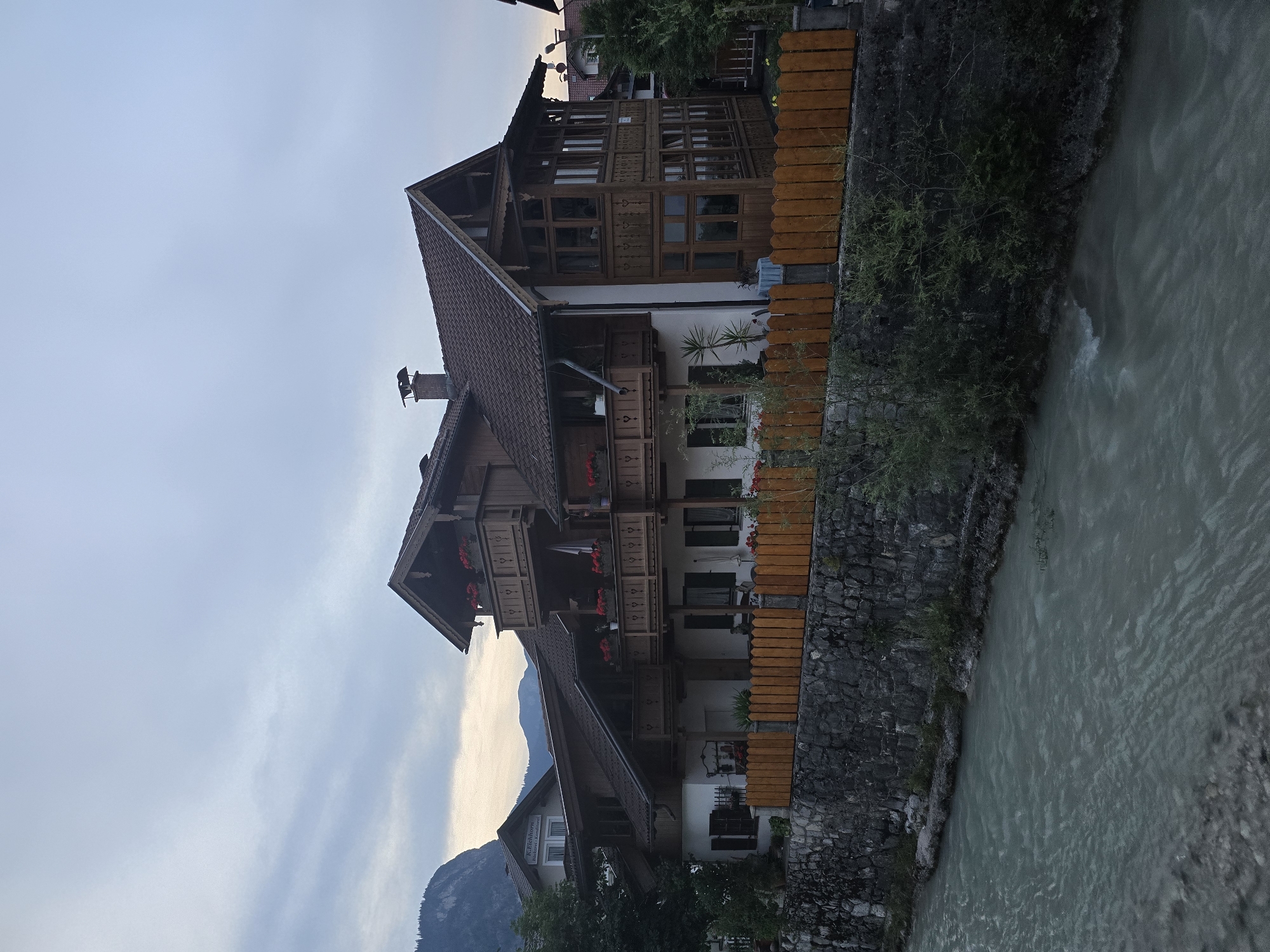 The image depicts a multi-story wooden house situated on a stone embankment overlooking a flowing river in Garmisch-Partenkirchen, Germany. The house features multiple balconies adorned with potted red flowers and intricate wooden carvings. A light brown wooden fence runs along the top of the embankment, separating the property from the river. The sky is overcast with muted tones, and the surrounding landscape includes trees and distant mountains. The scene appears to be captured during daylight, likely in the late afternoon or early evening given the soft lighting. No people are visible in the image.