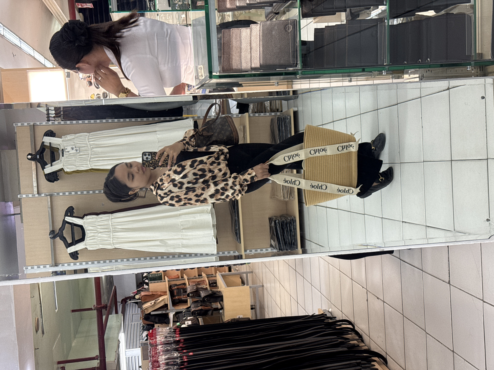 The image captures a reflection in a large mirror within a retail store. A young woman with her hair in a bun is taking a selfie with her phone, wearing a leopard print blouse and black pants. She is carrying a woven straw bag with "CHLOE" written on the straps. In the background, through the mirror, clothing racks with dresses and shelves stocked with handbags and wallets are visible. The floor is tiled. The reflection also shows another woman wearing a white shirt and glasses in the upper right portion of the image, looking towards the woman taking the selfie. The retail environment appears to be in a mall setting. Based on the context of "CHLOE" branding and the general upscale appearance of the merchandise, this scene is likely within a department store or a high-end boutique in a mall located in Davao City, Philippines.