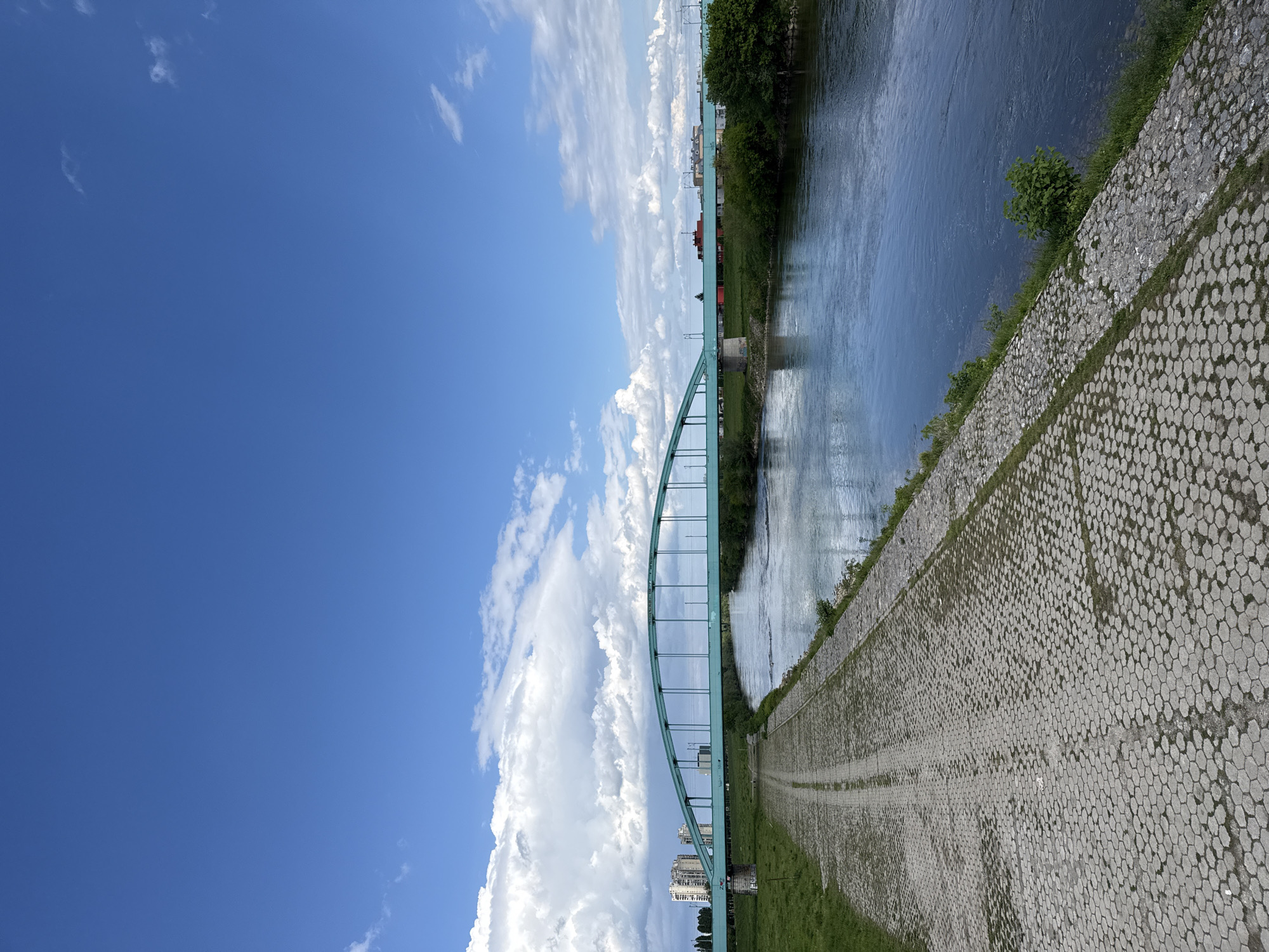The image presents a serene daytime landscape featuring a river, a distinctive arch bridge, and a vast blue sky. A wide river flows across the scene, its surface reflecting the sky and creating subtle patterns.
Spanning the river is a prominent arch bridge, distinguished by its light green or turquoise color. The bridge features an arch-through-deck design, with vertical elements supporting the roadway from the main arch structure. Along the near bank, a meticulously constructed embankment is visible, composed of numerous hexagonal concrete blocks. Patches of green grass grow between these blocks and along the top edge of the embankment, which also appears to have a pathway running alongside the river.
The far bank of the river is bordered by a line of dense green trees and vegetation. Beyond these trees, a glimpse of an urban environment with various buildings can be seen in the distance. Overhead lines, likely power or tram lines, are visible above the bridge on the distant side.
The sky, occupying the upper left portion of the image, is a clear, vibrant blue, dotted with scattered white cumulus clouds. The bright and even lighting suggests a sunny day, likely during the late morning or early afternoon. No people or visible text are present in the image.
This scene captures a peaceful moment along the Sava River in Zagreb, Croatia, with the iconic Hendrik Jordan Bridge (Zeleni Most) serving as a central architectural element within the urban landscape.