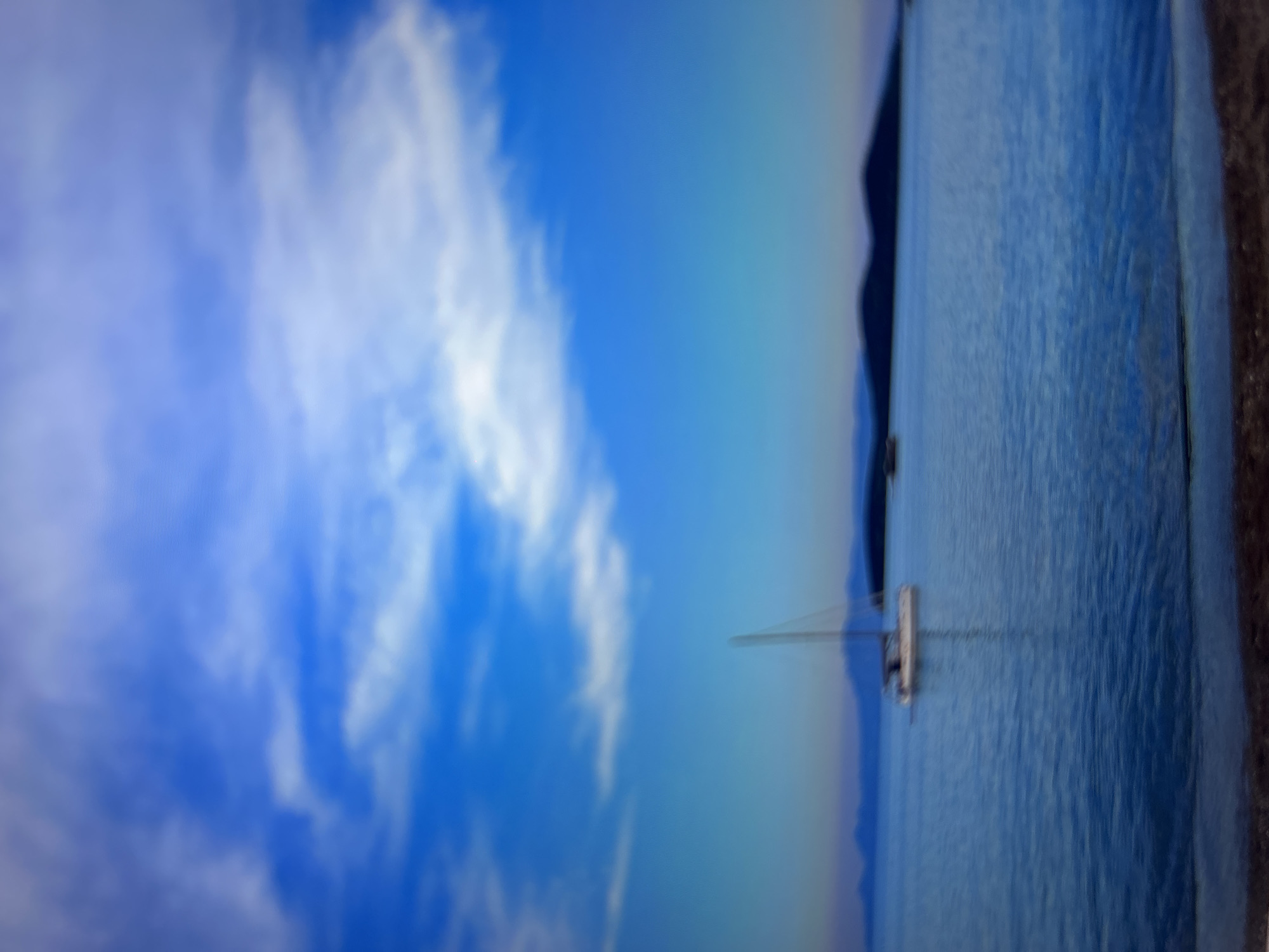 The image shows a view of a body of water, possibly a bay or harbor, with a sailboat on the water's surface. The sky above is blue with white, puffy clouds. In the distance, there is a landmass or coastline visible. The image seems to have been taken at an angle, as if the viewer is looking out from an elevated position. There is no indication of Sofia, Bulgaria, from the image.