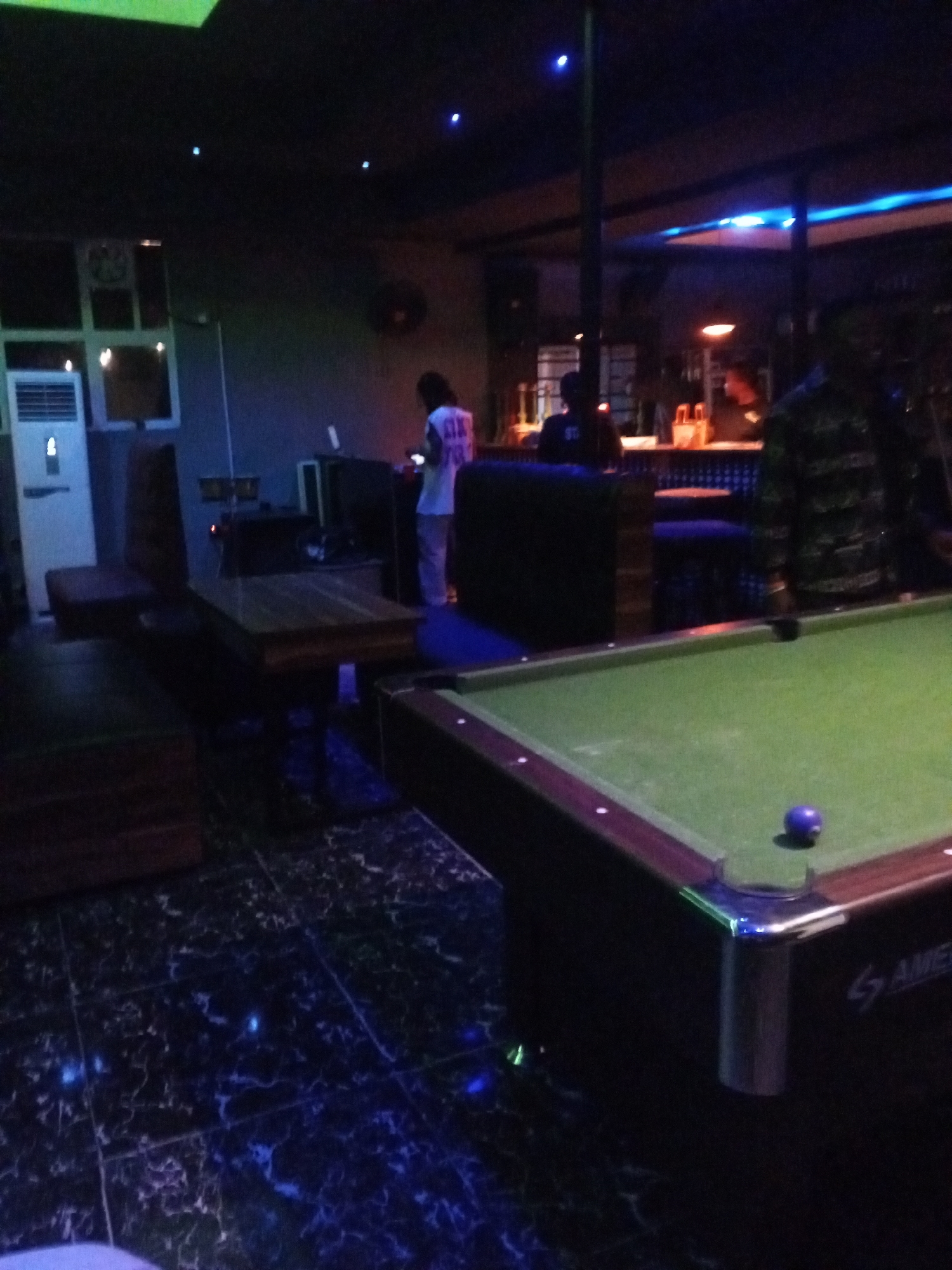 The image depicts the interior of what appears to be a recreational establishment, possibly a bar or lounge, in Lokoja, Nigeria. In the foreground, a green billiard table with a single purple ball is visible. To the left of the billiard table, a wooden table with cushioned seating is set up. Further back, an individual wearing a white t-shirt and light-colored pants stands near a counter, seemingly engaged with something on their phone. Another individual, dressed in a dark, patterned shirt, is partially visible on the right side of the image, facing away from the camera. The environment is dimly lit, with blue and green ambient lighting contributing to a relaxed atmosphere. A white air conditioning unit is positioned against a grey wall on the left side of the frame, and the floor has a dark, marbled tile pattern. The overall impression is that of a social gathering or leisure spot.
