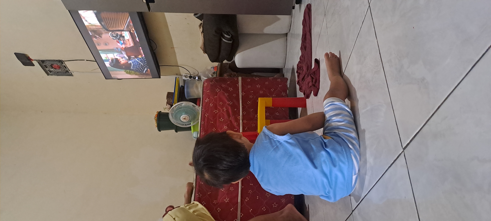 A young boy, wearing a light blue sleeveless shirt and striped shorts, sits on a tiled floor facing away from the camera, watching television. The television screen displays a scene with two people. Behind the boy, a red mattress with a star pattern is visible, with a small red and yellow toy structure on top of it. To the right of the mattress, a white stool and a folded dark red blanket are on the floor. In the background, a stack of dark green containers and a white oscillating fan are placed on top of the mattress. The room appears to be dimly lit, with light reflecting on the tiled floor. This image was taken in a residential area of Cemandi, Indonesia.