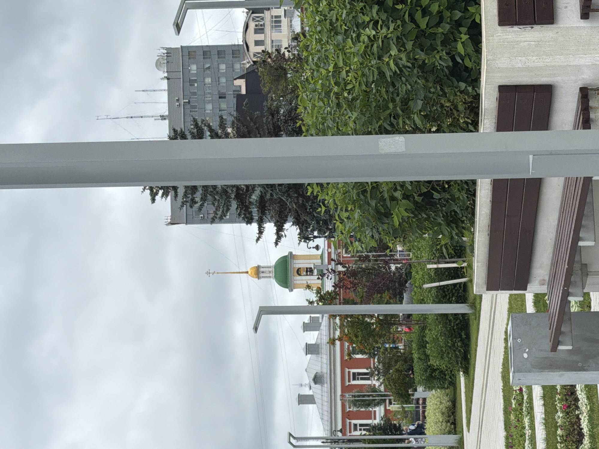 A medium-shot image, captured outdoors on an overcast day, shows a street scene in Perm, Russia, near the Perm Academic Theatre-Museum. In the foreground, a light grey, rectangular pillar stands vertically on the right side of the frame, obscuring a portion of the background. To the left of the pillar, a concrete bench with dark wooden slats is visible, with a portion of another similar bench to its right. Behind the bench, a concrete planter box and a trash receptacle are present.
Further back, a park-like area features neatly trimmed green bushes and some trees with autumnal foliage. In the mid-ground, a red building with white trim and several windows is visible, adjacent to a green-domed building topped with a golden cross. Behind these buildings, a taller, grey, multi-story structure with numerous windows and a flat roof with antennae stands out. The sky is heavily clouded, suggesting a dull or rainy day. A few indistinct figures can be seen walking in the distance, near the red building. The ground is paved with light-colored stone tiles, forming pathways.