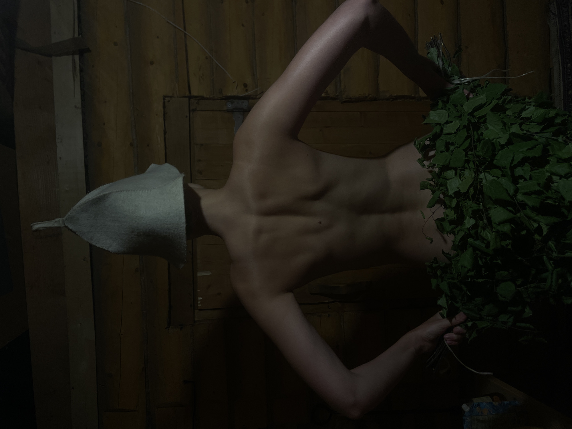 The image depicts a person viewed from the back, standing in a dimly lit, rustic wooden interior, identified as being in Sharkan, Russia. The individual wears a light grey felt bath hat, covering their head and neck. Their upper back, shoulders, and arms are bare, revealing a slender physique. From the lower back down, a dense arrangement of fresh green leafy branches, resembling birch *venik* used in banyas, acts as a covering, with the person's hands mostly obscured by the foliage at the sides. The background consists of horizontal wooden logs and planks, featuring a simple wooden door with visible hinges on the right side. A thin white cord stretches horizontally across the upper right wall. The overall lighting is low-key, highlighting parts of the skin while leaving much of the environment in soft shadow.