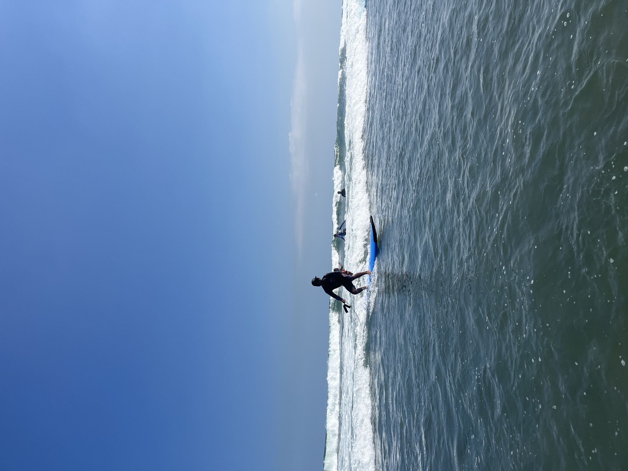 The image shows a surfing lesson on a bright, clear day at the beach in North Kuta, Indonesia. A lone surfer, dressed in dark attire and a cap, is in the foreground, standing on a blue surfboard and paddling through the waves. The surfer is holding a paddle, indicating they may be practicing stand-up paddleboarding or learning to surf. In the background, several other individuals on surfboards are visible further out in the ocean, also participating in surfing activities. The ocean is choppy with moderate waves breaking, and the sky is a vibrant, cloudless blue. The scene is captured from a low vantage point, at sea level, looking towards the shore.
