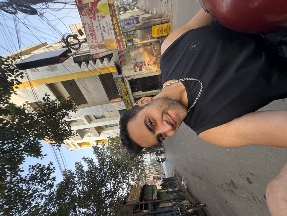 A man smiles at the camera while sitting on a motorcycle wearing a black tank top and silver chain. Buildings line a street in the background, and a sign with "Merry Christmas And Happy New Year 2026" is visible. Utility wires crisscross overhead, and trees are on the left side. The image is rotated counterclockwise.