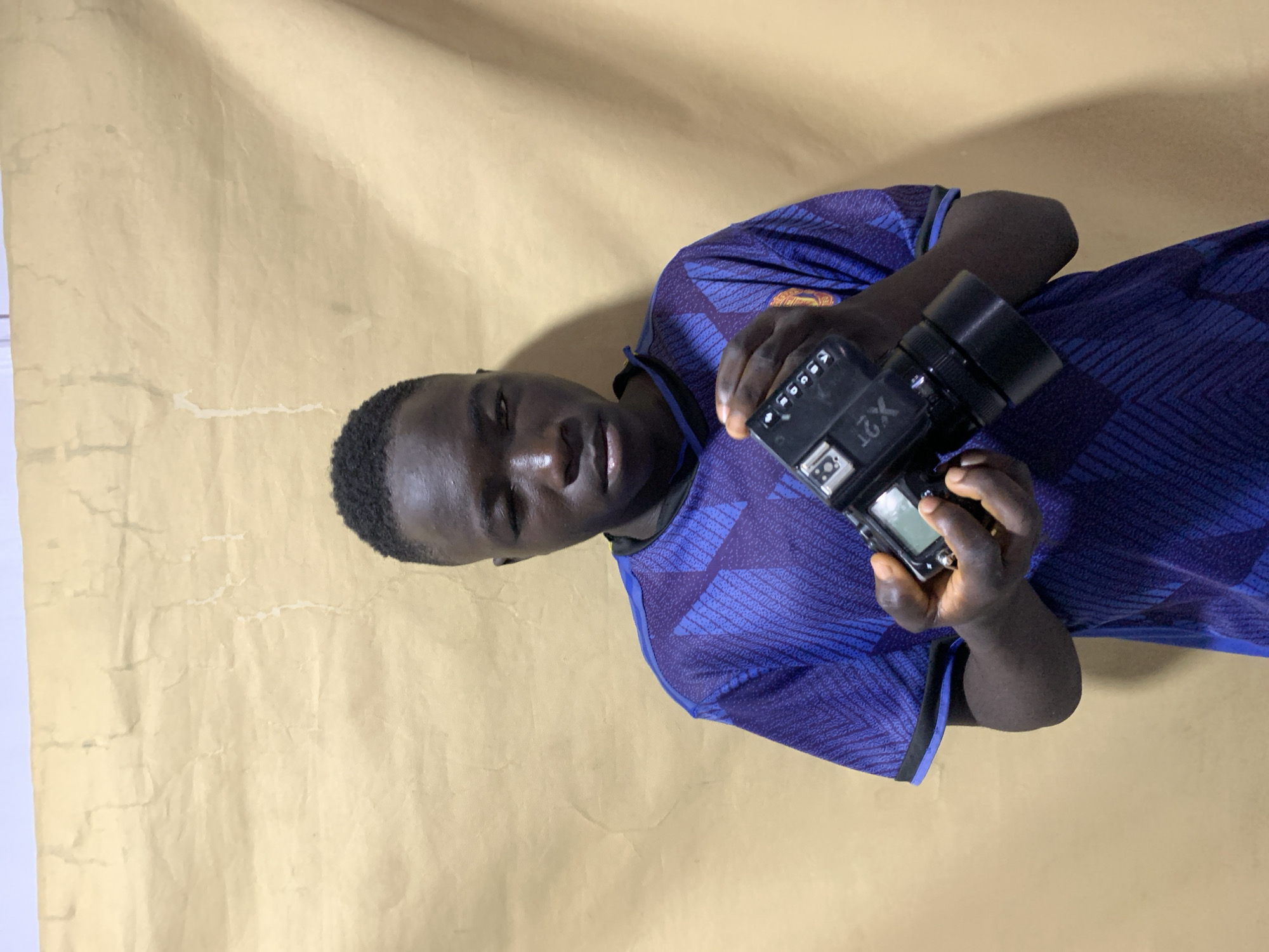 A young Black male, wearing a dark blue jersey with a geometric pattern, is holding a black camera. The background is a textured, light-brown sheet or backdrop. The location is Gura Top, Nigeria. The subject is looking at the camera he holds.