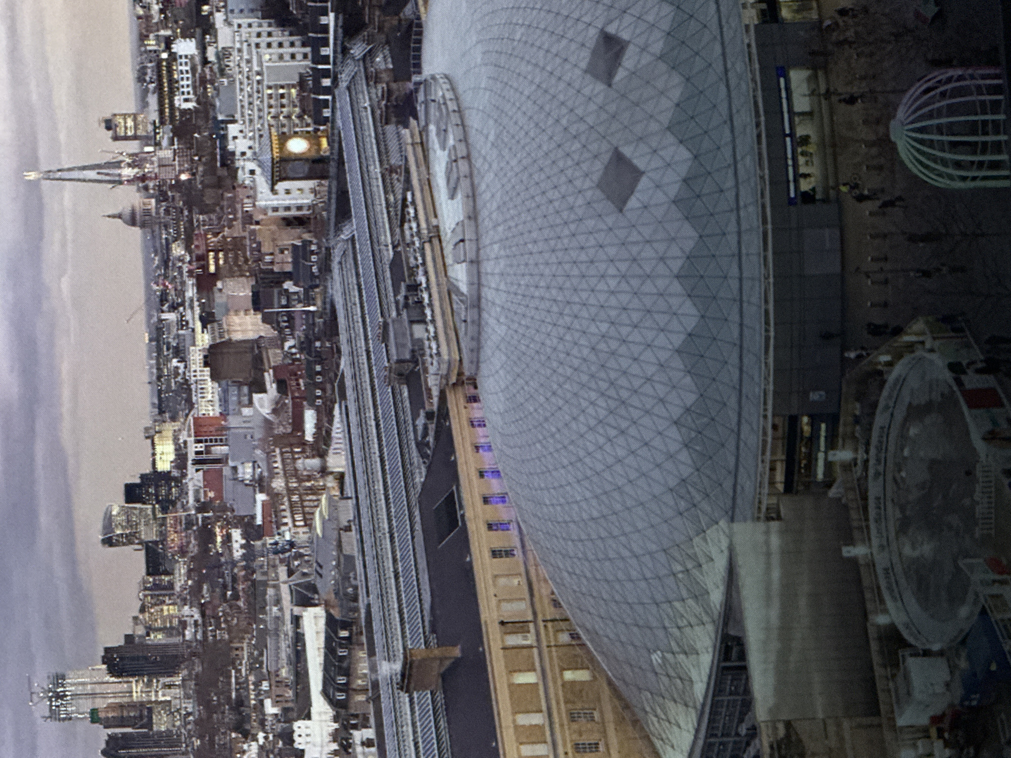 The image depicts a panoramic view of a city skyline, likely taken at dusk due to the muted lighting and cloudy sky. In the foreground, a large, modern glass-domed structure is prominent, characterized by its intricate geometric pattern. Adjacent to this dome is a long, beige building with numerous windows, some of which are illuminated with a purple light. Above these structures, a complex network of train tracks and platforms is visible, suggesting the presence of a major transportation hub.
The background reveals a dense urban landscape filled with various buildings, including towering skyscrapers and older, lower-rise structures. Notable landmarks in the distance include a tall, spire-like skyscraper and a domed building resembling a cathedral or religious institution. The overall impression is that of a bustling city, possibly undergoing development or renovation, given the visible construction elements in some parts of the skyline.
The image does not provide any information or visual cues that would suggest the location is Saratov, Russia. The architecture and urban planning visible are not characteristic of Saratov.
