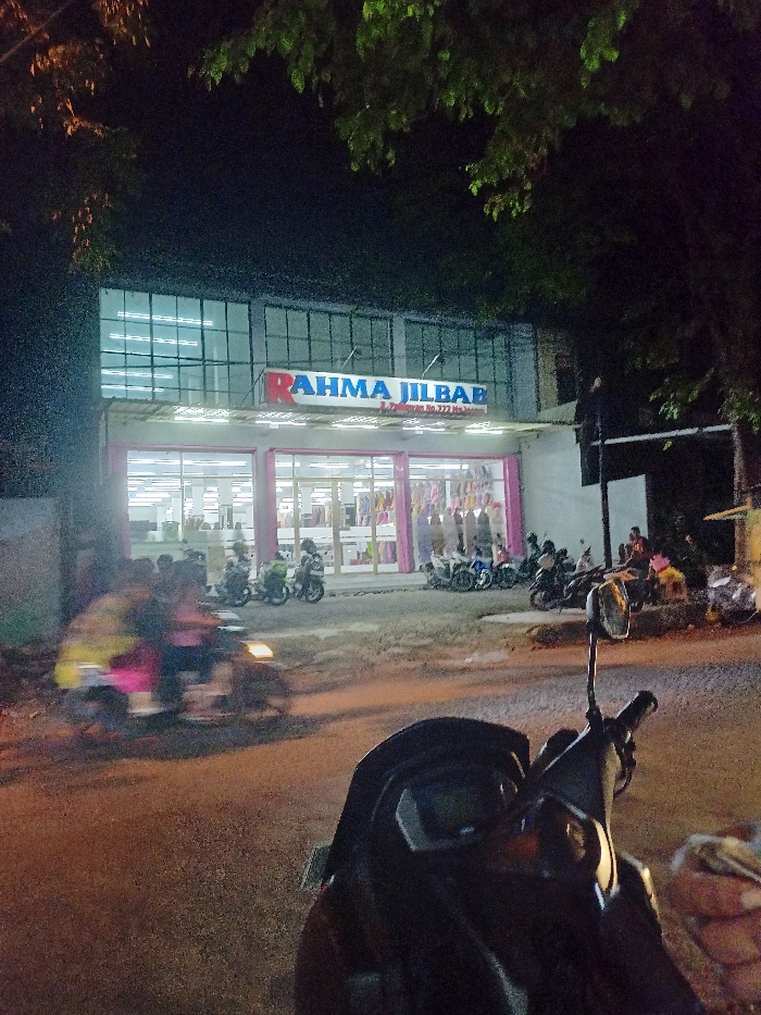 The image captures a nighttime street scene in Mojosari, Indonesia, dominated by a brightly lit store named "RAHMA JILBAB." The two-story building features large windows on both levels, with the ground floor acting as the main retail space, showcasing various garments, likely jilbabs, through its glass facade. The interior of the store appears brightly lit with fluorescent lights, revealing racks of clothing.
Above the ground floor entrance, a prominent red and blue sign reads "RAHMA JILBAB," with smaller text underneath indicating the address: "Jl. Pahlawan No. 222 Mojosari." The exterior of the store is well-illuminated, contrasting with the dark sky and silhouetted tree foliage overhead, suggesting a bustling evening atmosphere.
Several motorcycles are parked along the street and sidewalk in front of the shop, indicating patronage. A group of people is visible near the entrance and to the right of the store, some appearing to be seated or gathered. In the immediate foreground, the handlebars and rearview mirror of another motorcycle are partially visible, suggesting the perspective of someone parked or waiting. A blurred motorcycle with a rider is seen moving past on the road from left to right, its headlights faintly visible, adding a dynamic element to the scene. The weather appears clear and calm. The overall impression is one of a local commercial area active after dark, with shoppers and passersby illuminated by the store's lights.