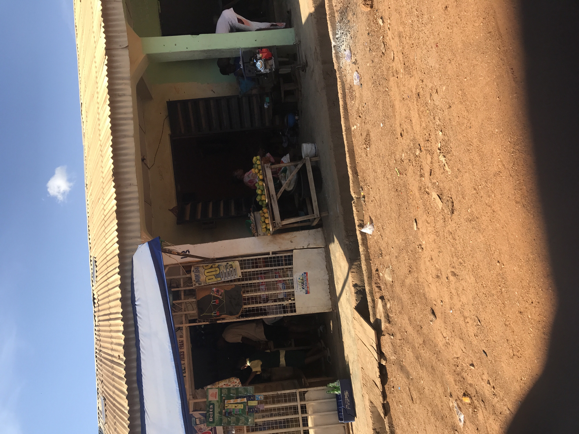 This media file captures a vibrant street scene in Ilorin, Nigeria, under bright daylight. The image is oriented vertically but depicts a horizontal stretch of an unpaved, reddish-brown dirt road next to a multi-story building housing small businesses.
On the left, the building features corrugated metal roofing, possibly on an overhang or upper floor, against a clear blue sky with a solitary white cloud. The building's facade is painted in muted tones, with a light green section on the upper floor and a lighter beige/yellow on the lower levels.
Several individuals are visible. On the upper floor balcony, a person in a white garment is seated. Below, another person is seen near a red and silver generator set, suggesting an off-grid power source. On the ground floor, a vendor is seated behind a wooden stand laden with yellow and green fruits, likely mangoes or oranges. Adjacent to this fruit stand is another shop, partially obscured by a metal gate or security bars. Inside, people are visible, and outside, crates of beverages, including "Delta" brand drinks and a Pepsi crate, are stacked. Various advertisements and signs are affixed to the shop front, with partially legible text including "POS" and "TEL 7033 946" and possibly "SNOW" or "SNOWS" written on the wall. A blue awning extends from one of the shops.
The road surface is dusty and uneven, with scattered small debris and rocks. A prominent dark shadow on the right side of the road suggests strong sunlight from a high angle, indicating it's likely midday. The overall scene portrays daily commerce and life in a bustling urban or semi-urban environment.