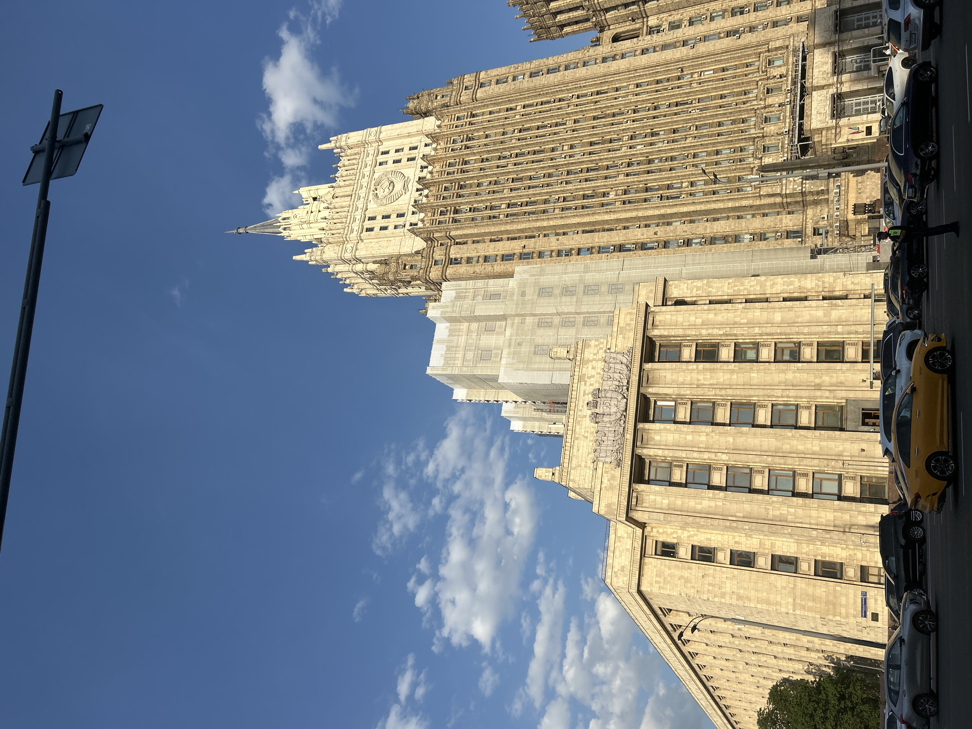 The image captures a view of the Ministry of Foreign Affairs building in Moscow, Russia, under a bright, partly cloudy sky. The building is a prominent example of Stalinist architecture, characterized by its imposing size, decorative elements, and distinct spire. Scaffolding partially covers one section of the building, indicating ongoing maintenance or renovation work. A row of parked cars is visible along the street in front of the building, including a yellow taxi. A street sign is present on the left side of the frame.