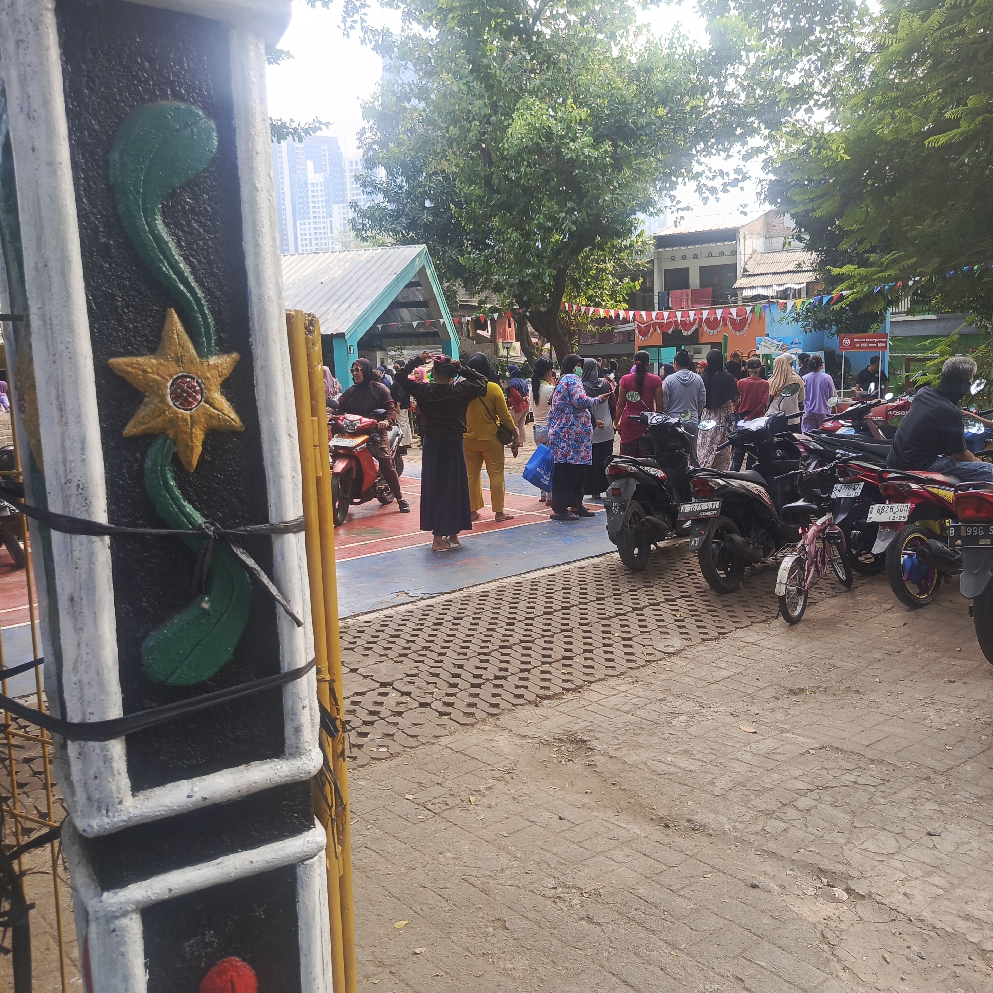 In South Jakarta, Indonesia, a bustling outdoor scene unfolds with a diverse group of people gathered near a building. A prominent pillar with a decorative star motif stands in the foreground, its textured surface contrasting with the surrounding environment. The ground is a mix of interlocking concrete blocks and paved surfaces, indicating an urban or semi-urban setting.
A variety of motorcycles are parked, suggesting a mode of transportation for many of the attendees. The people are dressed in casual attire, with many wearing headscarves, common in Indonesia. Some individuals are interacting with each other, while others appear to be observing or participating in an event.
The background reveals a simple, open-sided building with a corrugated metal roof, possibly a community hall or event space. Beyond this, trees provide a natural canopy, and hints of taller urban structures can be seen in the distance, characteristic of a city environment. Decorative banners, some featuring red and white patterns, add a festive touch to the scene, possibly indicating a celebration or local event. The overall atmosphere appears lively and communal.