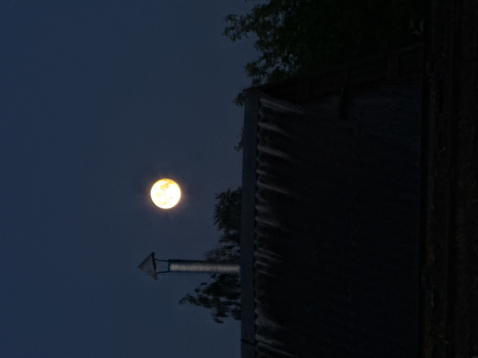 The image, oriented vertically, captures a moonlit night scene. A large, bright, yellowish-orange full moon dominates the left side of the frame against a dark, deep blue night sky, which shows some visible grain. On the right side, a dark, silhouetted structure, likely a building or shed, occupies a significant portion of the frame. Its dark silhouette reveals a textured surface, possibly corrugated metal or weathered wood, catching minimal light.
Protruding from the roofline of this structure and extending towards the moon is a distinct, light-colored metallic vent or small chimney with an angular, arrow-like cap. Dark, indistinct silhouettes of tree branches and foliage are visible behind and partially obscuring the structure and also closer to the vent, adding to the nighttime ambiance. The scene is static, devoid of human activity or visible text. The overall impression is one of a quiet, peaceful night, likely in a rural or semi-rural setting, consistent with the given location context of Лабжанскі сельскі Савет, Belarus.
