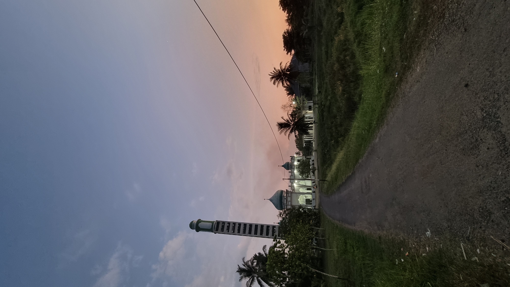 In Teupinsiron, Indonesia, at dusk, a dirt path leads towards a mosque with a prominent, slender minaret. The mosque has a turquoise dome and is illuminated, with visible lights in its windows. Coconut trees and other vegetation surround the building. The sky transitions from a pale blue at the top to shades of pink and orange near the horizon, indicating the setting sun. A power line stretches diagonally across the upper right portion of the image. The foreground features the textured surface of the path and grassy verges, with some scattered debris on the left side. No people are visible in the scene. The overall atmosphere is tranquil and serene.