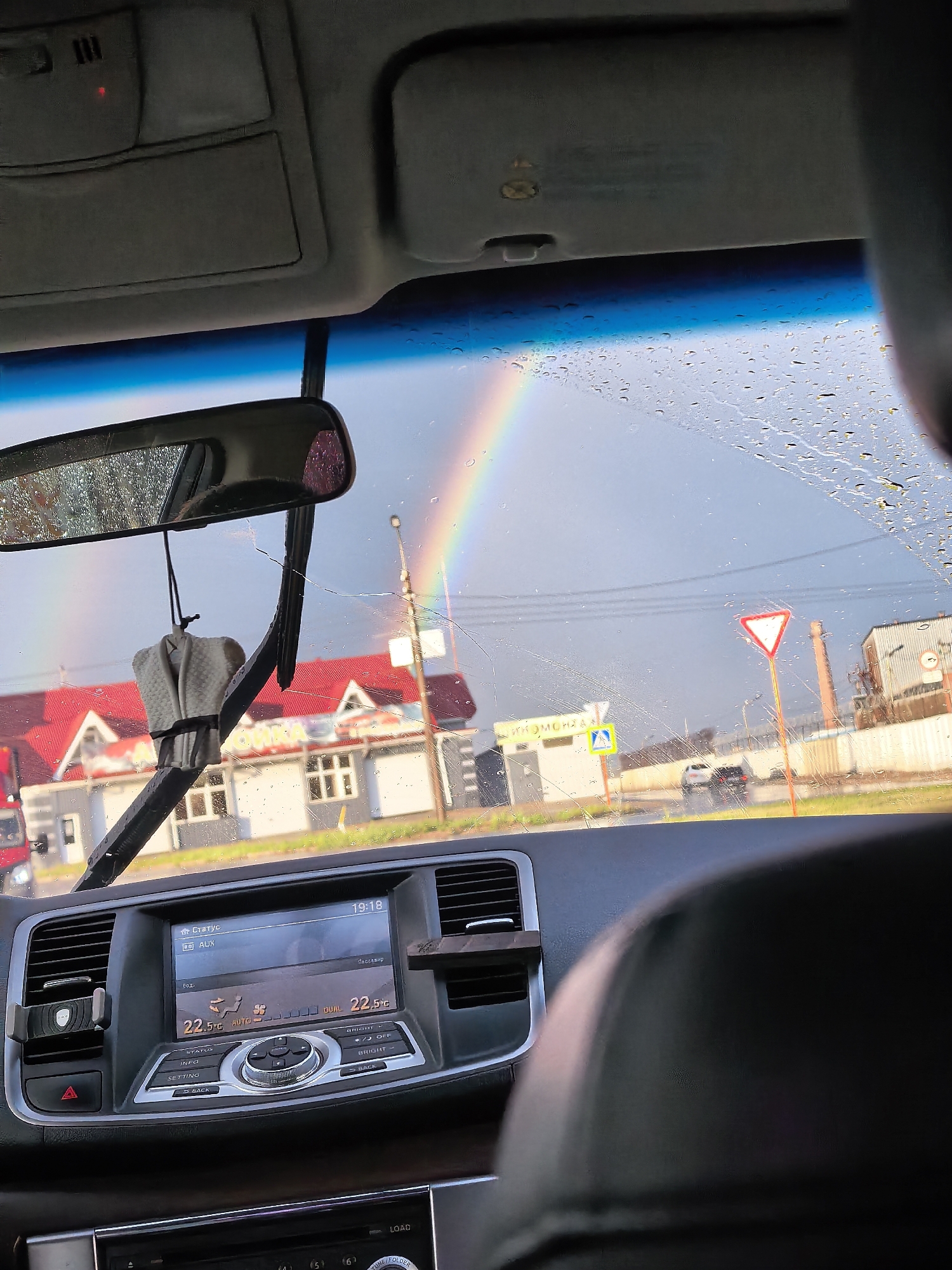 The image, taken from inside a car in Sterlitamak, Russia, captures a scene with a rainbow visible through a rain-streaked windshield. The car's interior shows the dashboard with a functioning climate control system displaying a temperature of 22.5°C. A car air freshener hangs from the rearview mirror. Outside, buildings with red roofs, industrial structures, and street signs are visible. The time displayed on the car's screen is 19:18. The overall environment suggests a post-rainy, possibly evening, setting in an urban or semi-urban area.