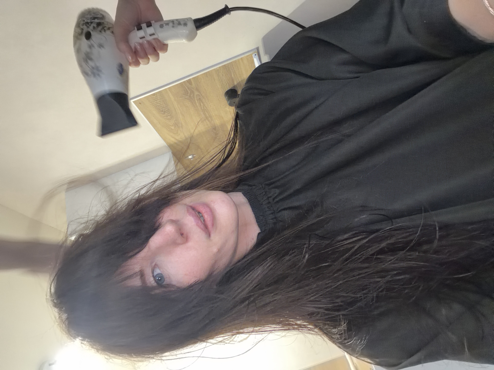 A person with long, dark hair and light skin is having their hair dried with a white and gray hair dryer. The person is wearing a black garment that covers their shoulders and chest. The background appears to be indoors with light-colored walls and a cabinet or countertop visible. The location in Perm, Russia, is not evident from the image.