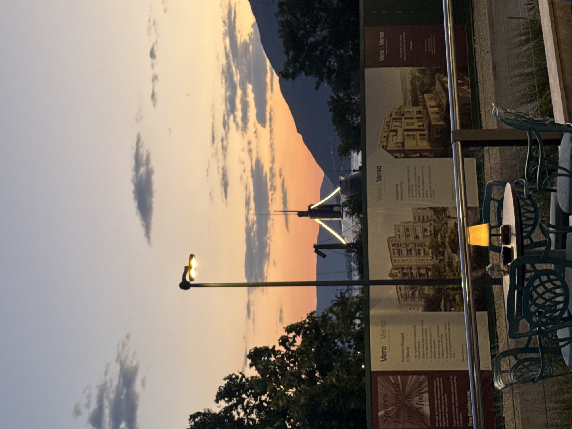 Here's a description of the image:
The image, taken at dusk or early evening, shows an outdoor setting in Tivat, Montenegro. The sky displays a blend of colors – light blue with some clouds transitioning to orange closer to the horizon, likely indicating sunset.
In the background, a mountain range is visible, silhouetted against the sky. A structure or building under construction, with illuminated scaffolding, stands out against the colored sky. A street lamp is positioned in the foreground, with its dual lights illuminated.
To the right of the frame, there's an outdoor seating area. A round table and several ornate, green metal chairs are visible. On the table, there are some objects, likely glasses. Behind the chairs, there's a fence or barrier with advertising boards showing images of a building project labelled "Vero Versa", possibly promoting real estate. The trees that frame the sky on the side of the image is a green tree.