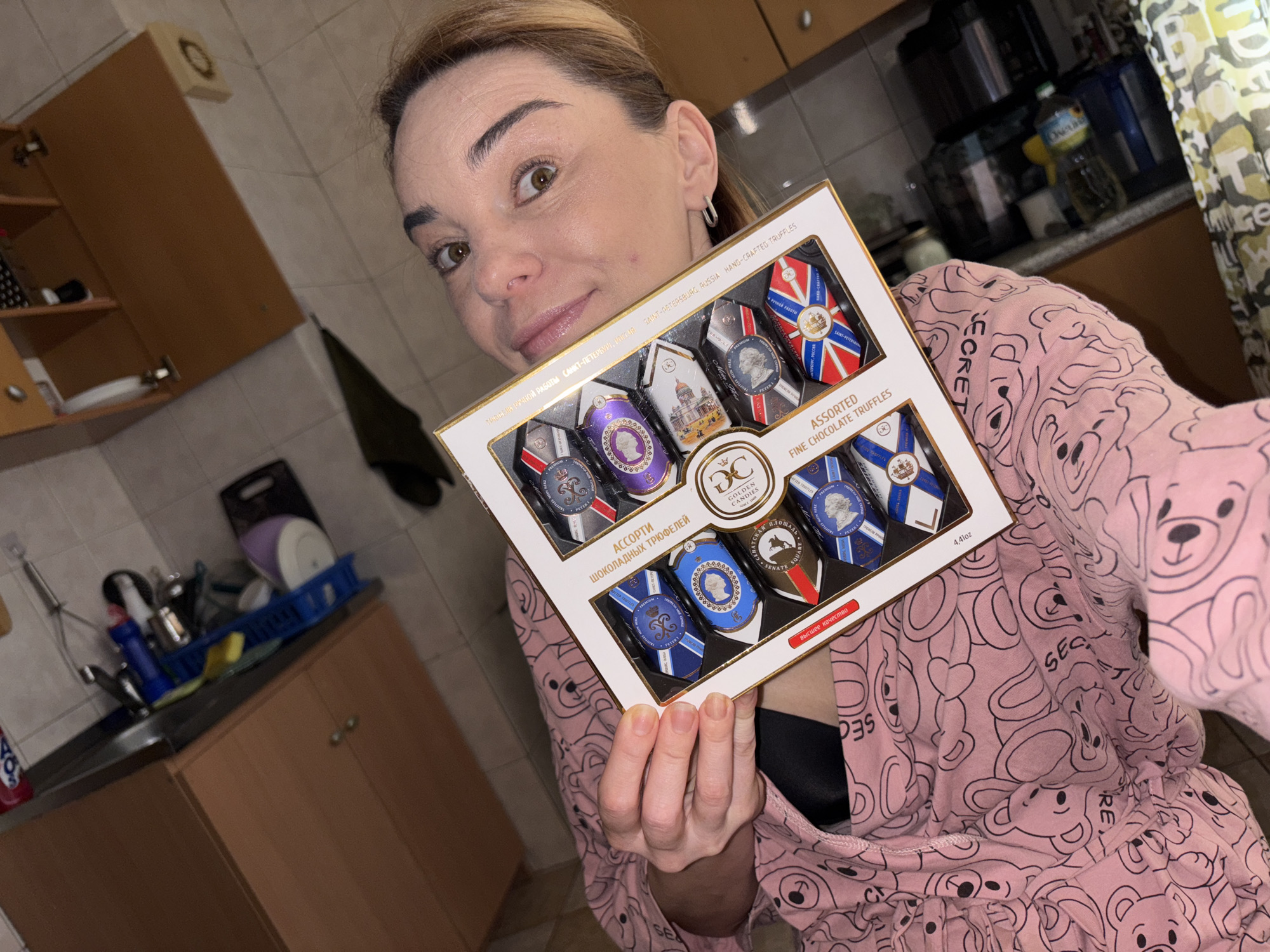 A woman, with blonde hair pulled back and wearing a pink robe with a teddy bear pattern, is holding a box of assorted fine chocolate truffles. The box is white with gold trim and features images of Saint Petersburg landmarks. The woman's face is close to the camera, and she has a surprised expression. The background shows a kitchen counter with a sink and cabinets. The scene is indoors.