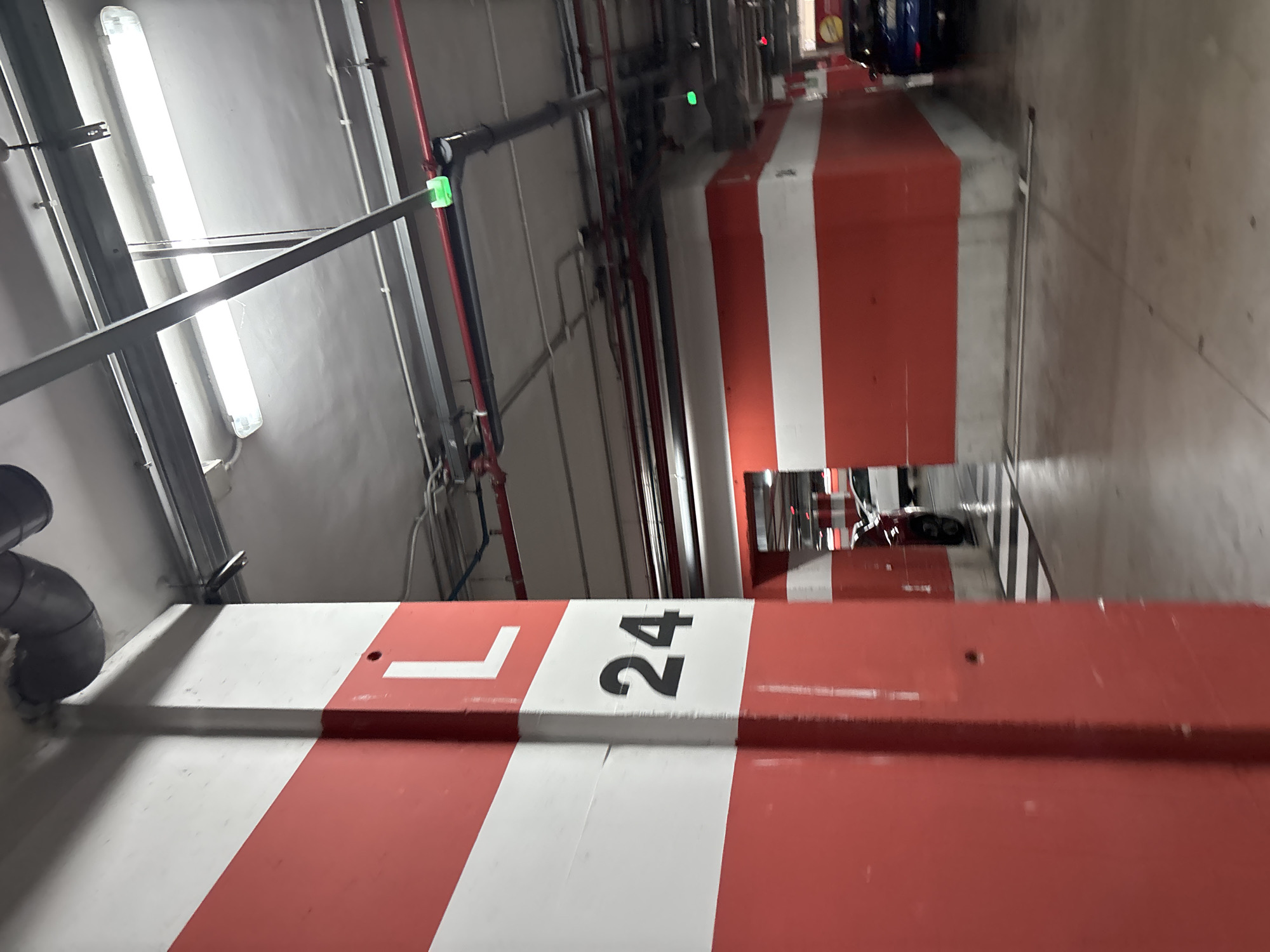 The image depicts a section of an indoor parking garage. A large, red and white striped concrete pillar in the foreground is marked with the letter "L" and the number "24". In the background, the garage features red and white striped walls, fluorescent lighting fixtures on the ceiling, and various pipes and conduits. Two cars are visible in the distance. The environment is well-lit and appears to be clean. There are no people visible in the image. The overall scene is that of a typical, functional parking facility.