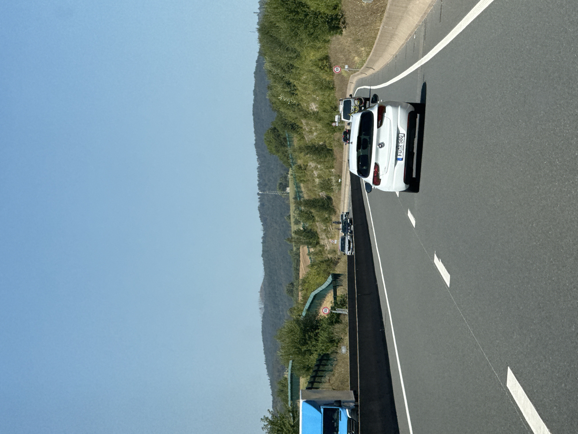 The image shows a section of a highway in or near Bad Lauterberg im Harz, Germany. The perspective is from a vehicle traveling on the highway. In the foreground, the asphalt road with white lane markings is visible. Several vehicles are on the road, including a white passenger car in the right lane, and a blue truck on the left. In the background, a wooded hillside with a clear blue sky above it is depicted. A speed limit sign indicating 100 km/h is visible on the right side of the road. The environment appears to be rural or semi-rural, with natural landscape surrounding the highway.