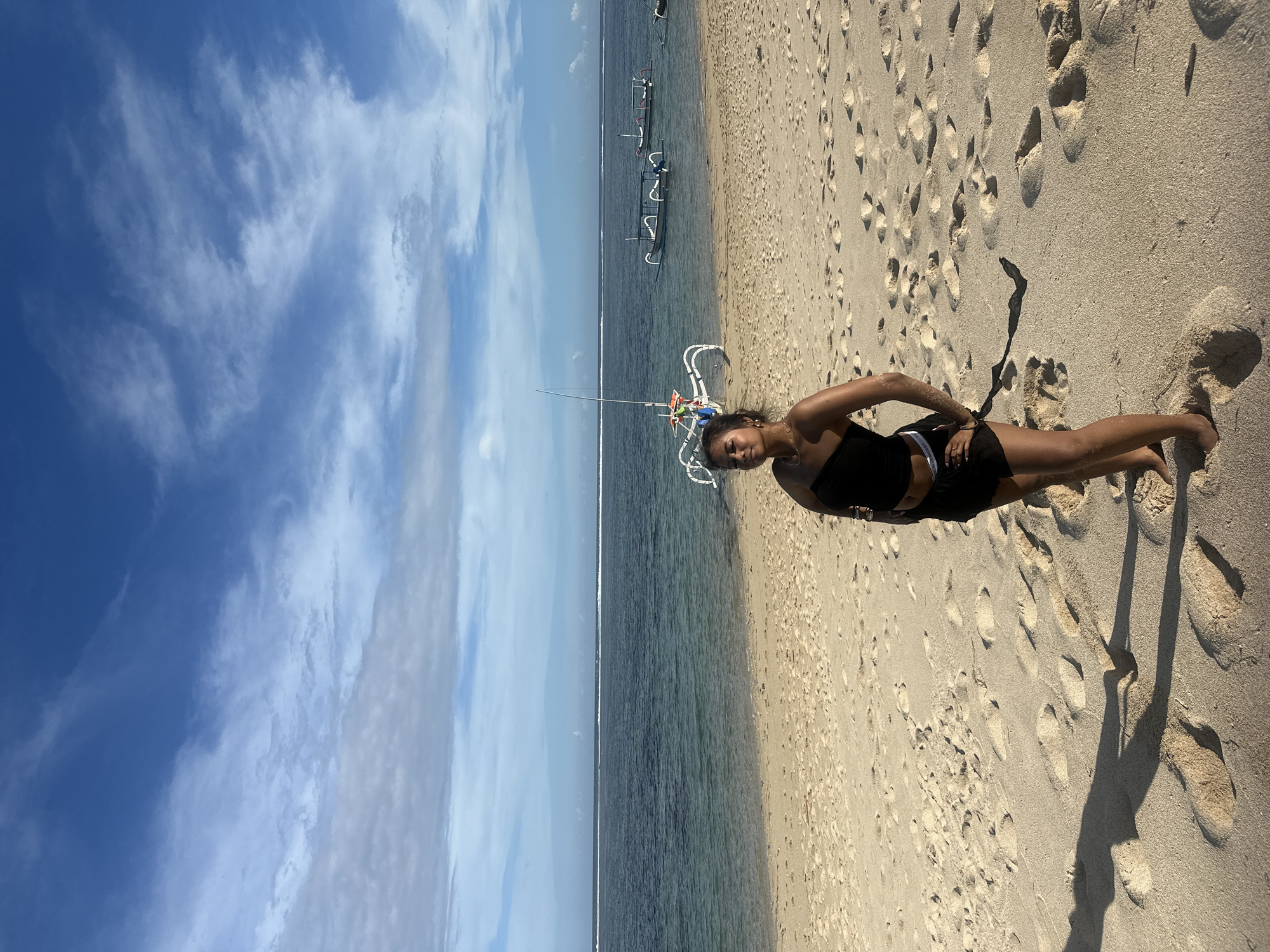 A young woman in a black top and shorts stands on a sandy beach in Kuta Selatan, Indonesia. The sky is light blue with scattered clouds. The ocean is a darker shade of blue, separating the sky and the beach. A few boats are visible in the distance. The sand is covered in footprints. The woman's shadow extends away from her on the beach.