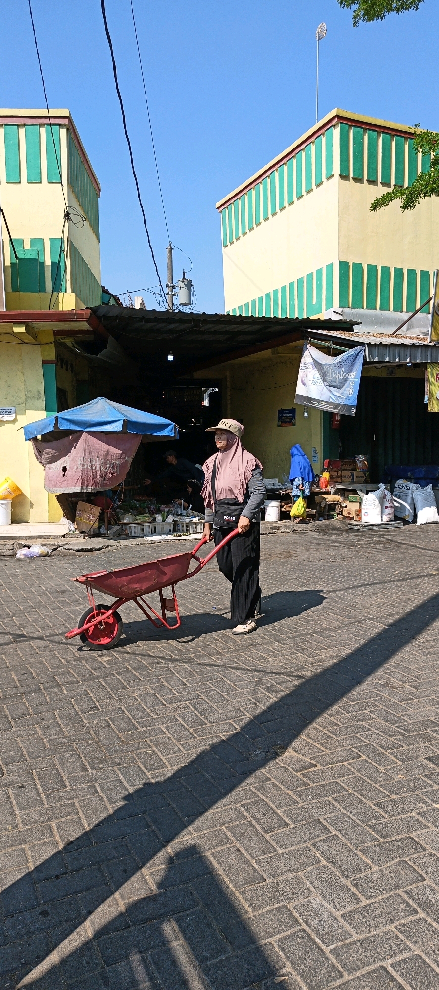 The image depicts a street scene in the city of Klaten, Indonesia, characterized by the presence of commercial buildings and a pedestrian. A woman, dressed in a pink hijab, black pants, and a grey top with a black bag bearing the word "POLLO", is pushing a red wheelbarrow along a brick-paved street. She is walking away from the camera, towards the right side of the frame. The background reveals a marketplace with various stalls, awnings, and bundled goods. The buildings flanking the street are painted yellow with green accents. The sky is clear and blue, indicating a sunny day, and long shadows are cast across the pavement, suggesting it is either morning or late afternoon. The overall impression is of a busy, working market environment within an urban setting.