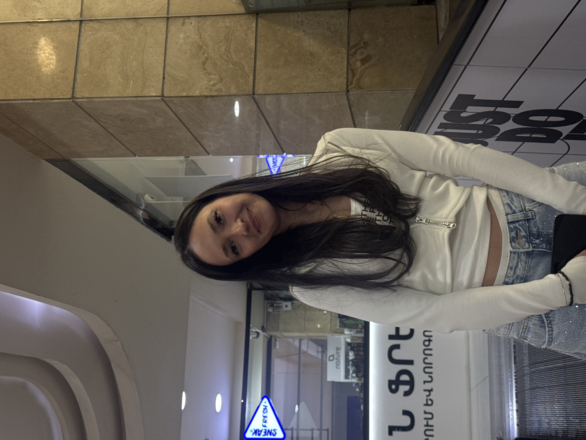 A young woman with dark hair is posing for a picture inside what appears to be a shopping mall in Yerevan, Armenia. She's wearing a white long-sleeved crop top and light-wash denim jeans with embellishments. In the background, signs are written in Armenian. One sign visible through the glass reads "Sneak" and "Fresh". Also visible through the glass is a storefront for a "nature" brand. A Nike "Just Do It" sign is present in the scene.