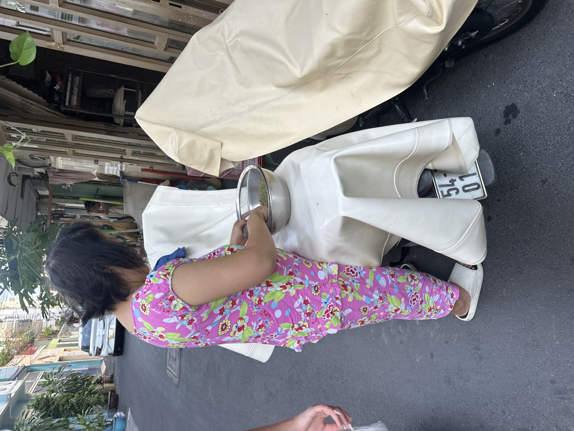 A woman in a pink floral pajama set and white sandals is shown from above, crouched and facing left while sieving green beans in a metal colander. She is positioned next to a white motorcycle seat, with a motorcycle cover partially visible behind it. The motorcycle's license plate reads "54-01." The scene is situated on a city street in Thủ Đức, Vietnam, as evidenced by the urban environment, including nearby buildings, trees, and a glimpse of traffic.