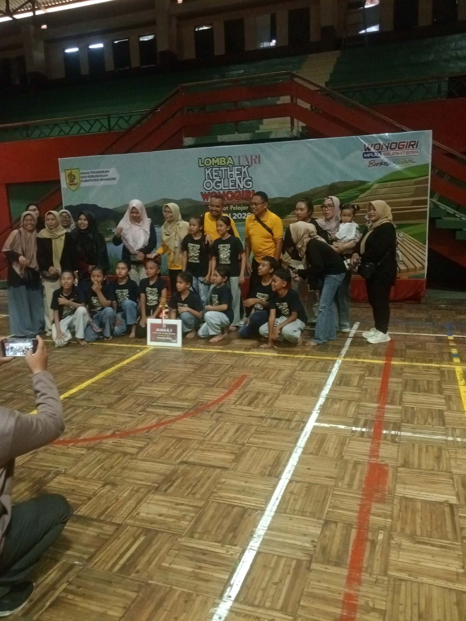 In Wuryorejo, Indonesia, a group of individuals, including children and adults, pose for a photograph on a wooden gymnasium floor. A banner in the background reads "LOMBA TARI KETHOK OGLENG WONOGIRI," indicating a traditional dance competition. Several children, dressed in matching black t-shirts, are seated in the front, some holding a trophy. Adults stand behind them, some in casual attire, others in headscarves. One person in the foreground is taking a photograph with a smartphone. The gymnasium features bleachers in the background and colored lines marking the court.