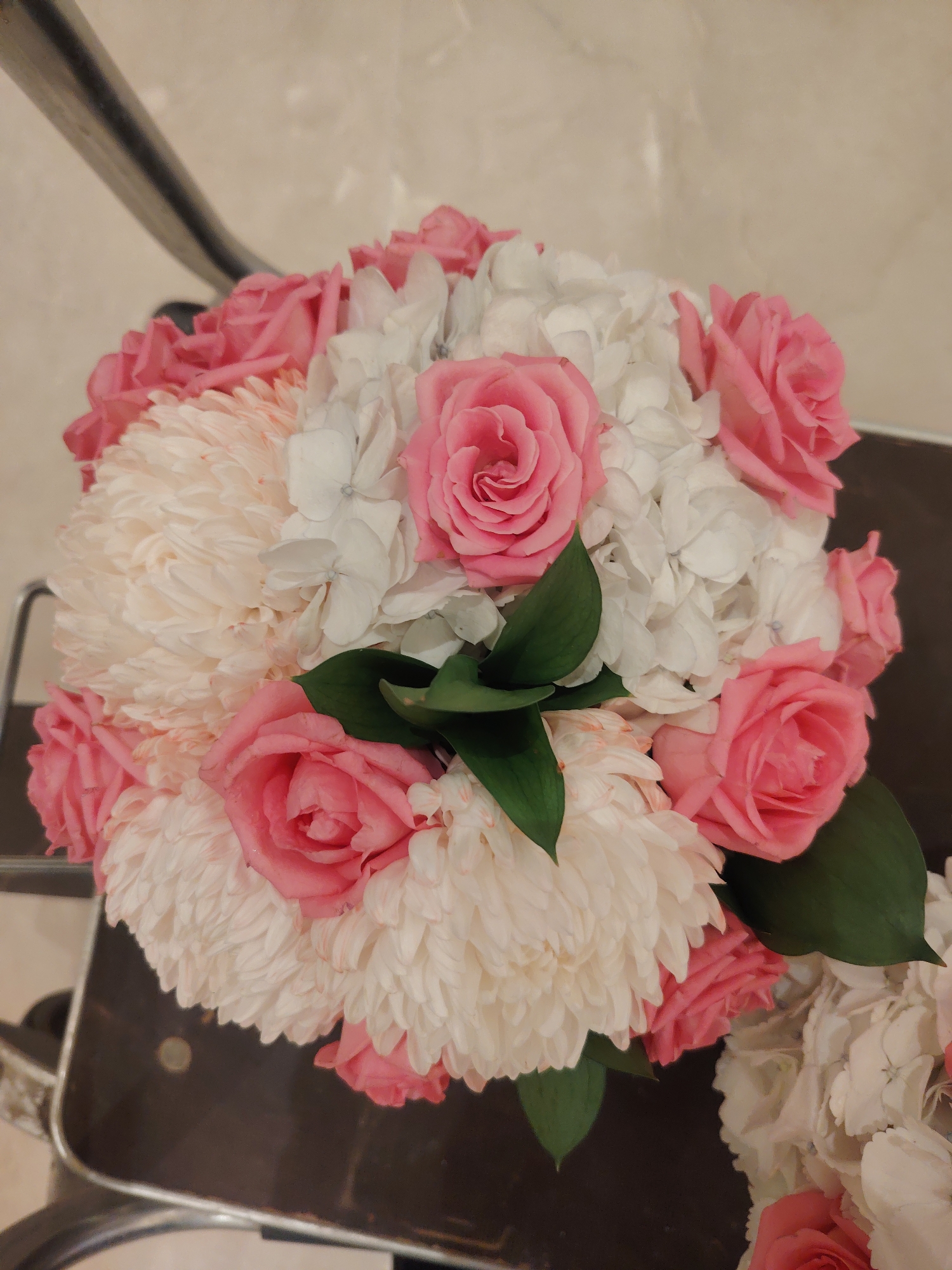 A bouquet of flowers, primarily pink roses and white hydrangeas with some white carnations, is arranged on a dark surface that appears to be part of a piece of furniture. The bouquet is the central focus of the image, with its various textures and colors clearly visible. The background is a neutral, light-colored wall or surface. The lighting suggests an indoor setting, possibly a room in a home or a venue. There are no people or animals in the image, and no specific landmarks in Konigil, Uzbekistan are identifiable.