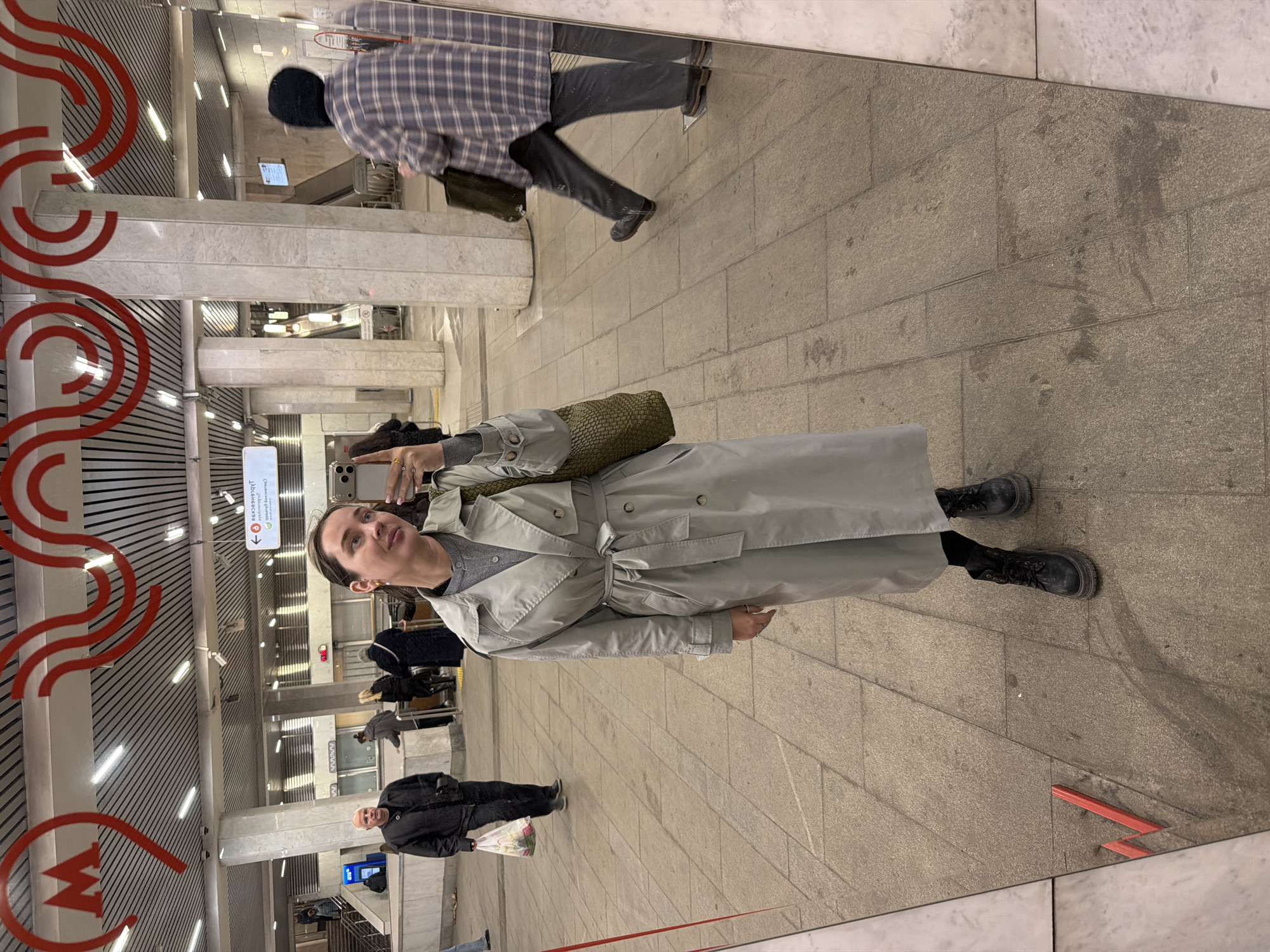 A full body selfie is taken in a mirror in the Moscow Metro. The woman in the photo is wearing a light colored trench coat, a gray sweater, and black boots. She is holding a phone in her right hand. In the background, other passengers are visible, and the Moscow Metro logo is displayed. The floor is made of rectangular gray tiles.