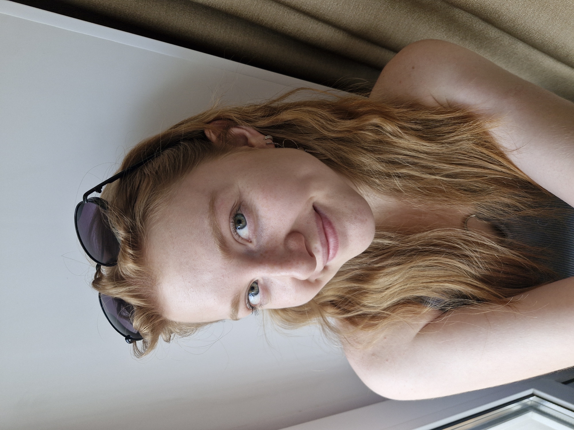 A young woman with long, strawberry-blonde, wavy hair is pictured indoors. She is fair-skinned, with green eyes, freckles, and light pink lips. Her hair is mostly down, with the front sections pulled back, holding a pair of dark sunglasses. She is wearing a gray tank top. The background consists of a white surface, which is likely a windowsill, and a tan curtain. The image is oriented at a tilted angle. The exact location is indeterminate as the image lacks any discernible landmarks to identify the setting as Yekaterinburg, Russia.