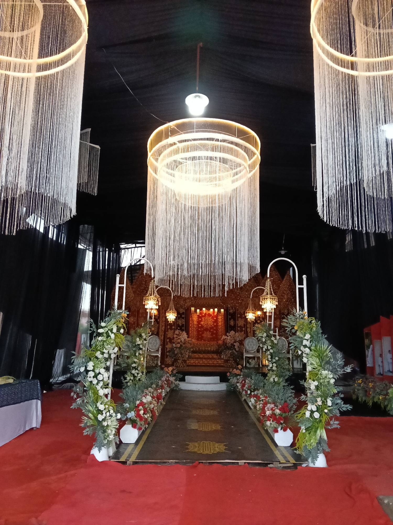 The image depicts an indoor wedding or event setup in Lhokseumawe, Indonesia. The focal point is a raised dais adorned with traditional Indonesian ornamentation and seating, illuminated by several candles and small chandeliers. A red carpet runs from the foreground to the dais, bordered by floral arrangements in white and red flowers. Above, large, round chandeliers with cascading white strands hang from the ceiling. The backdrop is black fabric, creating a darkened environment around the illuminated stage. The venue appears to be spacious, possibly a hall or tented area.