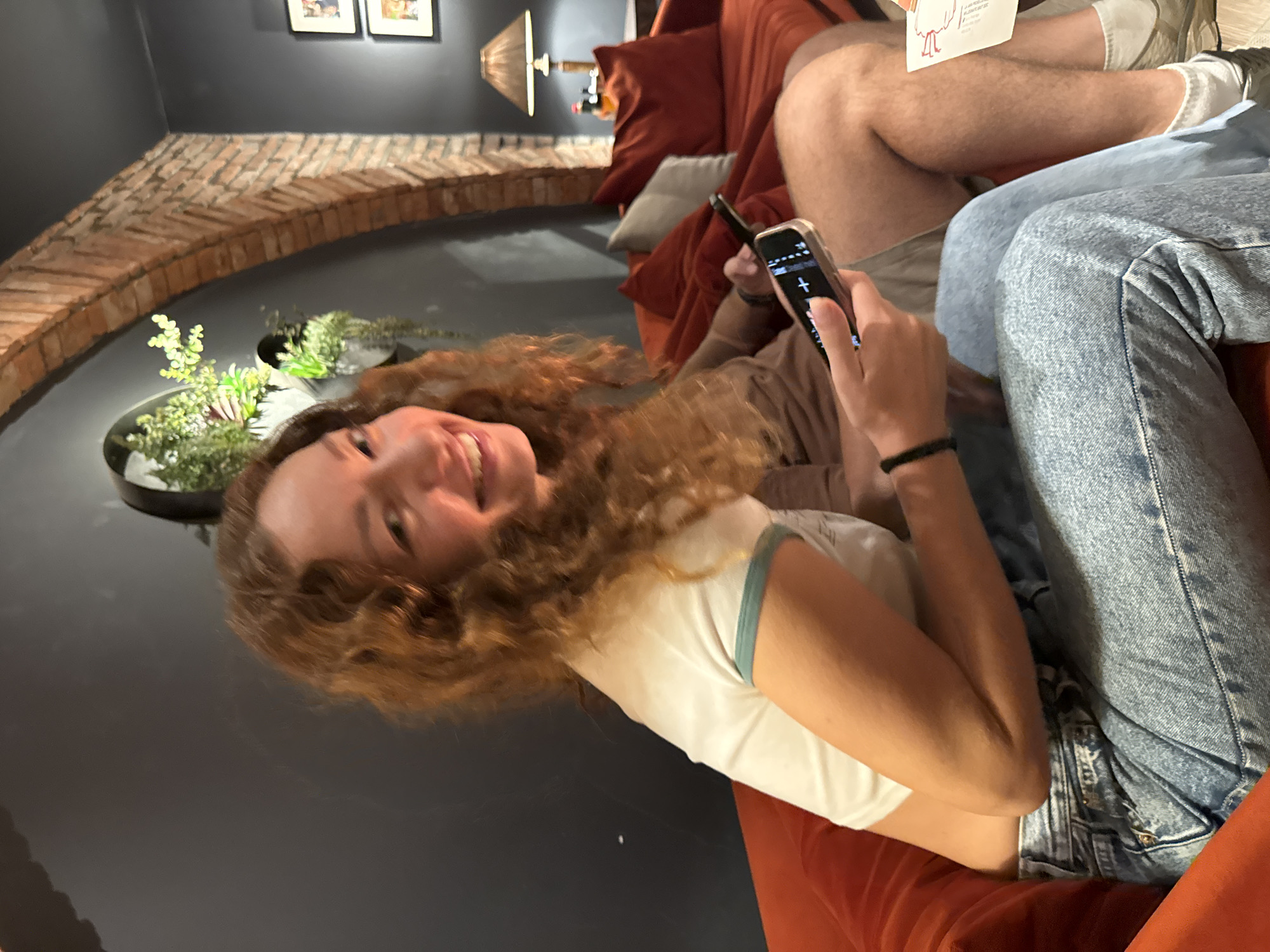 The image shows a young woman lying down on an orange couch, smiling and looking at the camera. She has long, wavy, reddish-brown hair and is wearing a white t-shirt with a green trim. She is holding a smartphone in her right hand, which displays a simple interface with a plus sign. Beside her, another person's legs are visible, wearing blue jeans.
The setting appears to be indoors, likely a living room or lounge area. The background features a dark gray wall with exposed brickwork forming an arch. There are some potted plants on a surface below the brickwork. On the wall above the brickwork, two framed pictures are hung. A lamp with a warm glow is visible in the background, suggesting it might be evening or the lighting is dim. The overall mood seems relaxed and casual, with the woman's smile conveying a sense of happiness or amusement. There is no visible text in the image except for what appears on the phone screen.