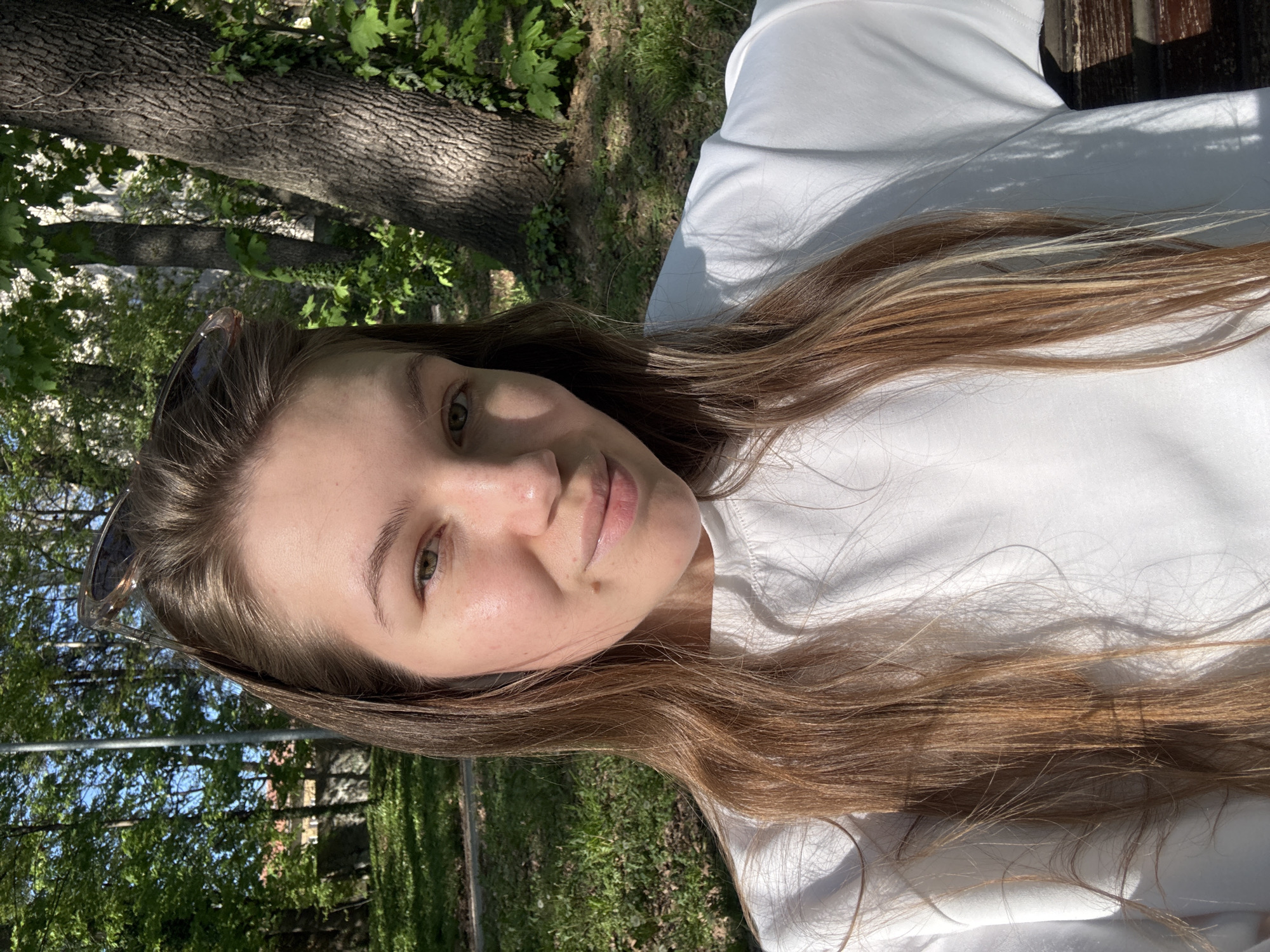A young woman with long, light brown hair is posing for a selfie outdoors. She is wearing a white shirt. The background features green grass, trees, and the building of Vozdovac Urban Municipality in Vozdovac, Serbia. Sunglasses are perched on her head. The image is taken from a low angle, looking up at her face.