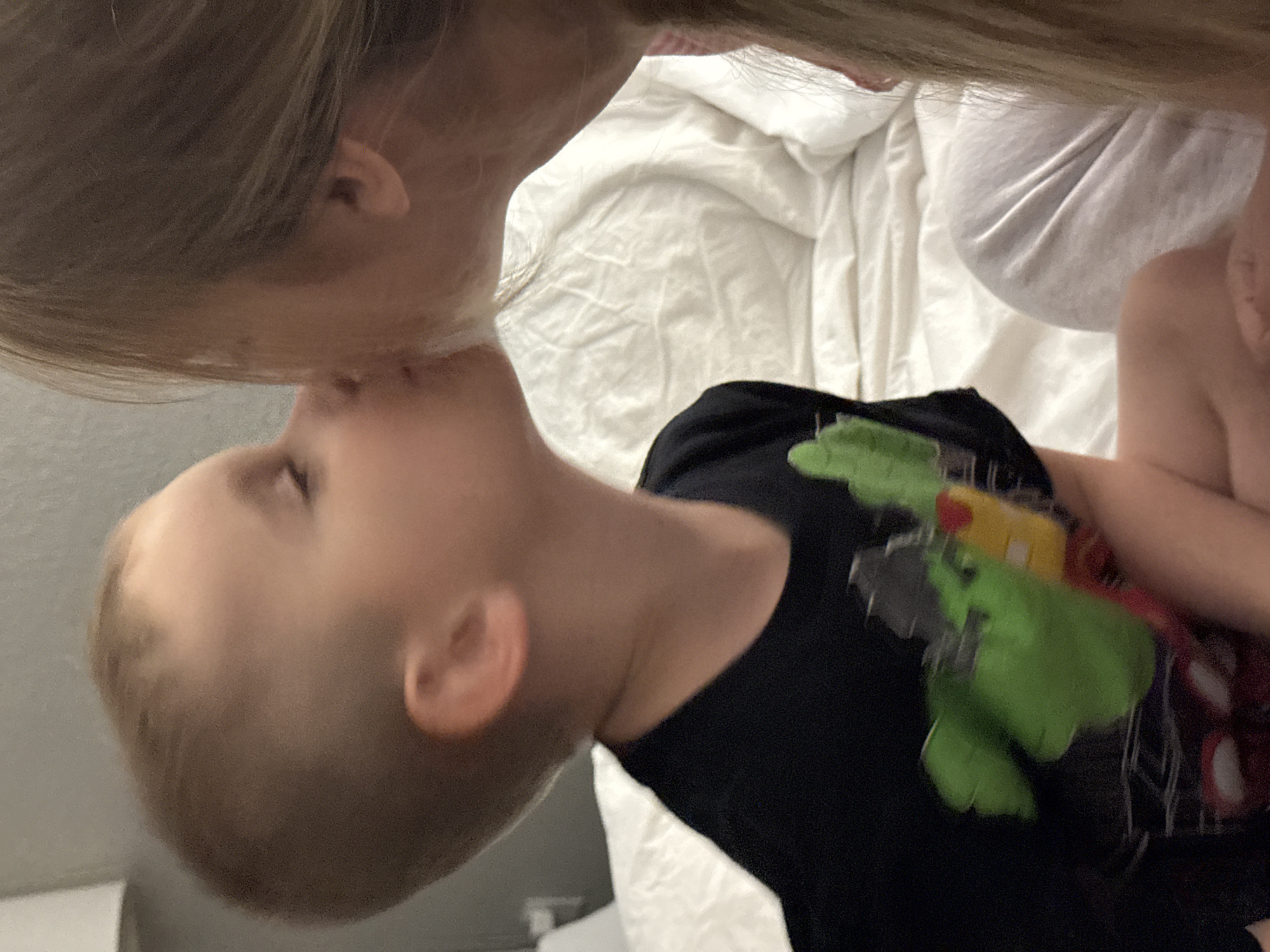 In this close-up image, a person with long blonde hair is leaning down to kiss a young child on the cheek. The child, with short blonde hair, is looking upwards, and their face is partially obscured by the adult's hair. The child is wearing a black t-shirt with a colorful graphic of what appears to be a cartoon character, possibly the Hulk, on it. The background consists of white bedding, suggesting an indoor setting, likely a bedroom. The lighting is soft, and the overall mood appears tender and affectionate. There is no visible text in the image. The context provided places this scene in Myrtle Beach, United States of America.