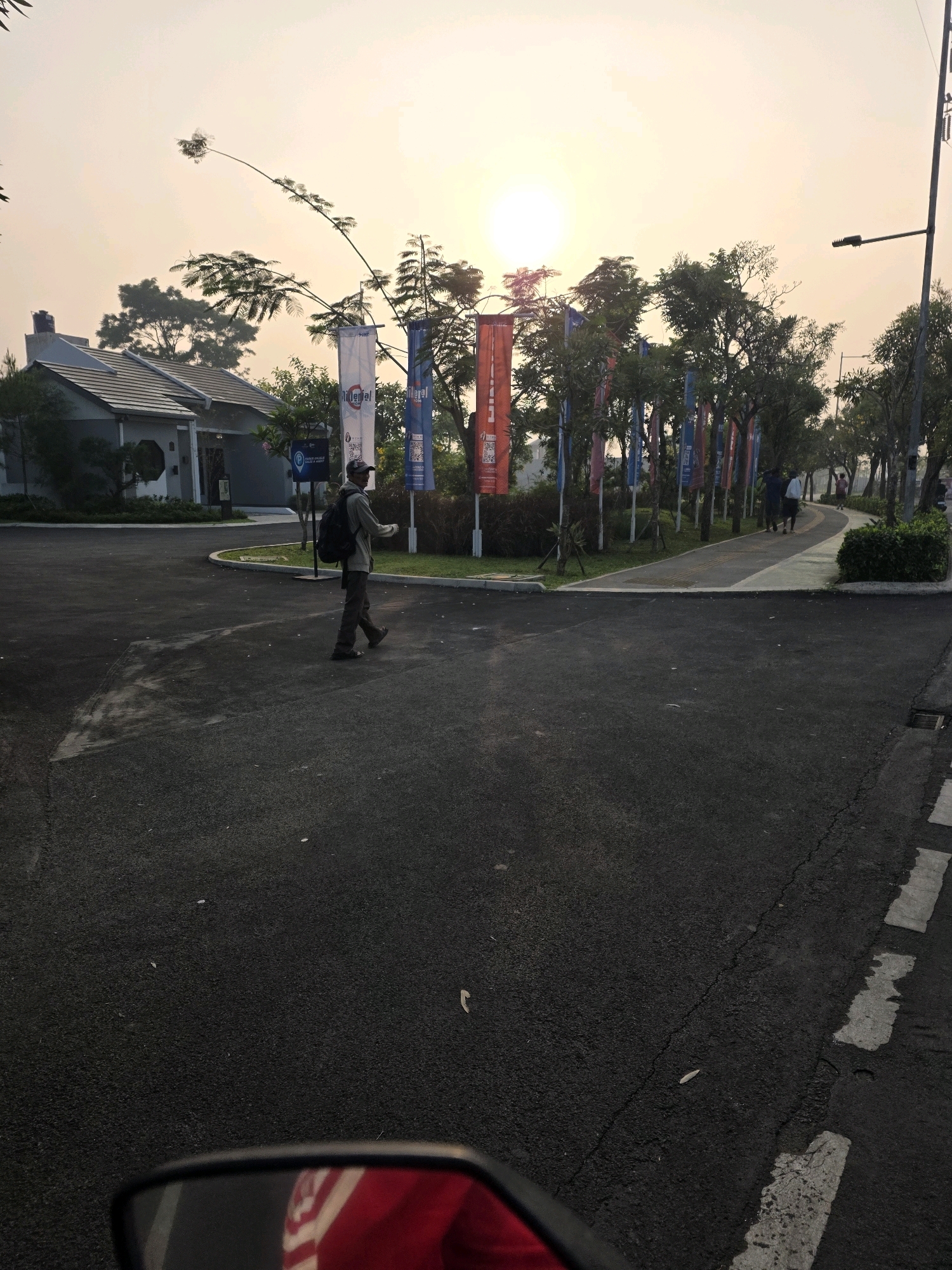 The image depicts a street scene in what appears to be a residential development in Bojongsoang, Indonesia. The foreground shows a paved road with white lane markings. In the middle ground, a person wearing a backpack and a cap is walking on the road. To the left, a modern house with a gray exterior and a tiled roof is visible. Beyond the house, several flags are planted along a pathway lined with trees. The flags are vertical and colorful, with some displaying text and graphics. The background features a bright sky with the sun setting or rising, casting a warm glow over the scene. Several other people can be seen walking on the pathway in the distance. The overall environment suggests a planned community with amenities such as walkways and decorative flags.