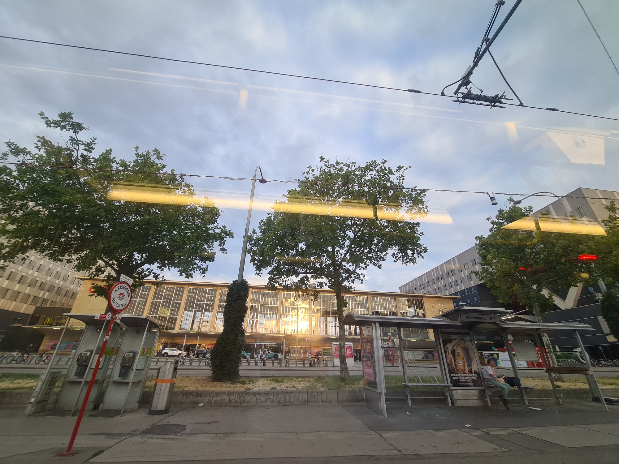 The image captures a view from inside a tram in Vienna, Austria, looking out at the Westbahnhof train station. The time of day appears to be late afternoon or early evening, with the sun reflecting brightly off the facade of the station building, which features large glass windows and a modern architectural style.
In the foreground, a tram stop shelter is visible on the right side of the frame. A person is seated on a bench within the shelter, looking to their left. Posters are displayed on the advertising panels of the shelter. To the left of the station, another tram stop area is visible with two vintage telephone booths and a red pole with signage. Trees with lush green foliage are interspersed throughout the scene, adding to the urban greenery.
Above the street level, overhead tram wires are suspended, with a trolley pole of the tram the viewer is on visible near the top of the image. The sky is partly cloudy, with soft light filtering through. The background shows the imposing structure of the Westbahnhof station and surrounding modern buildings. Signs indicate the location as "Westbahnhof" and "STRASSENBAHN HALTESTELLE" (tram stop). Bicycles are parked in racks in front of the station. The overall impression is of a busy urban transit hub on a pleasant day.