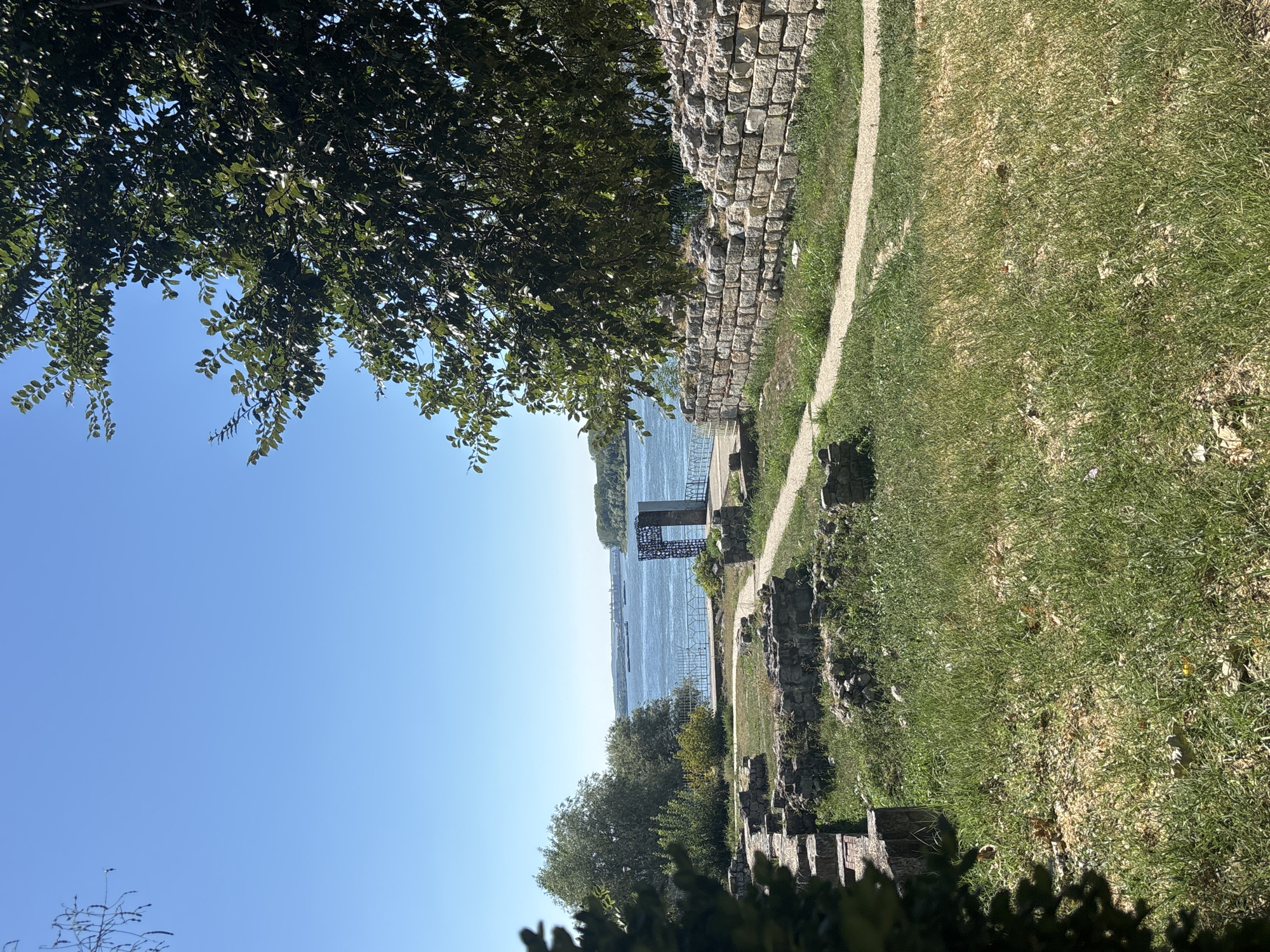 The image captures a sunny outdoor scene with a clear blue sky. In the foreground, there are the ruins of stone structures overgrown with grass. A dirt path winds through the ruins and leads towards a stone wall on the right side of the image.
Beyond the ruins, a wide body of water, likely the Danube River, is visible. Several barges are seen on the water in the distance. On the far bank of the river, a tree line and some buildings are present.
In the middle ground, to the right of the river, there is a modern, abstract metal sculpture or monument. A fence runs along the edge of the embankment near the river. The overall environment suggests an ancient historical site preserved with modern additions, located in the city of Silistra, Bulgaria, known for its Roman heritage and its location on the Danube River. The scene is devoid of any people.