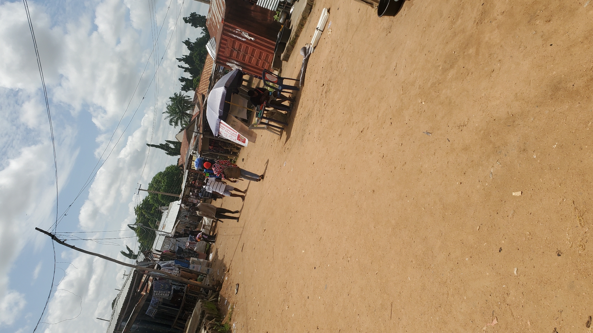 The image depicts a bustling marketplace in Jalingo, Nigeria. The ground is covered in reddish-brown dirt, indicating an outdoor setting. On the left side of the frame, there are several market stalls with corrugated metal roofs, displaying various goods, including clothing and textiles. A wooden pole with tangled wires extends upwards from the stalls.
In the center of the image, several individuals are walking along the dirt path. One person, wearing a light-colored shirt and dark pants, is walking away from the camera. Another person, in a striped shirt and shorts, is walking in the same direction. A third person, with a red cap and a backpack, is walking towards the right side of the frame.
On the right side of the image, a market stall is set up under a large umbrella. A person is seated at a table beneath the umbrella, appearing to be engaged in an activity. Two blue plastic chairs are placed around the table. Adjacent to the stall is a large, red shipping container that appears to be used as part of the market. Further to the right, there are black plastic containers and a stack of white pipes. The sky is partly cloudy with scattered white clouds against a blue sky. The overall scene suggests a typical day in a Nigerian marketplace.