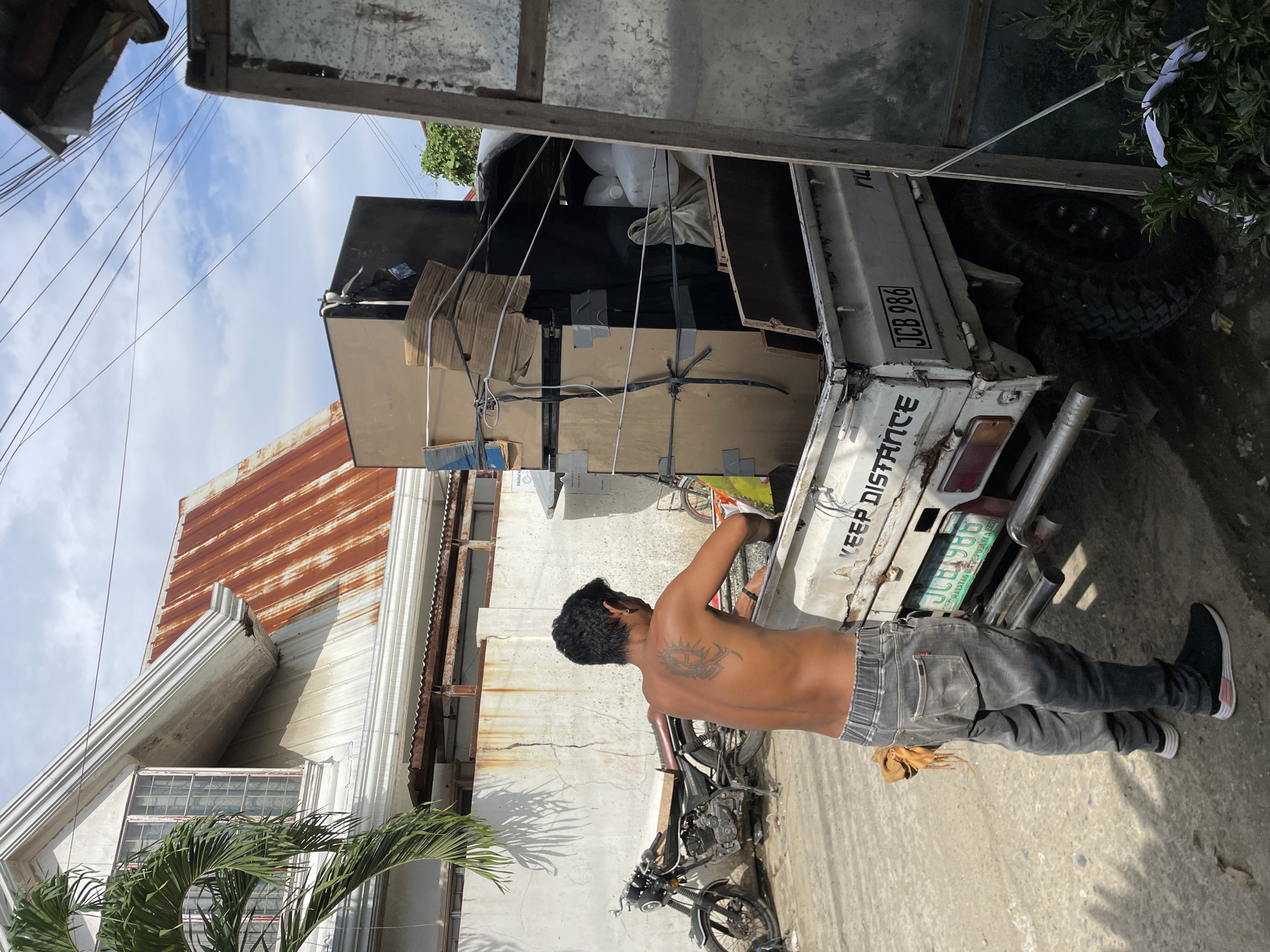 This image captures a man interacting with a heavily loaded white utility truck on a sunny day in what appears to be Davao City, Philippines.
The central subject is a shirtless man, seen from behind, with dark hair and a prominent tattoo on his upper back, resembling a tribal or sun-like design. He wears grey jeans with an elastic waistband and black sneakers with white soles. He is standing at the rear of the truck, seemingly engaged in securing or arranging the cargo.
The white truck is the primary object, packed with various items, including cardboard boxes, wooden or particle board panels, and several white bags or containers, suggesting a moving or delivery operation. The cargo is secured with ropes and straps. A black motorcycle is parked on the ground near the man, partially in shadow.
In the background, there's a building with a rusty corrugated iron roof and a white facade featuring ornate architectural trim. Another simpler white wall is also visible. Palm fronds extend into the frame from the foreground, and utility lines crisscross against the bright, blue sky with scattered white clouds. The harsh shadows indicate it's midday or early afternoon.
Visible text on the truck includes "KEEP DISTANCE" on the tailgate. Below it, a green license plate displays "JCB 986", with smaller text that appears to read "DAVAO", "PHILIPPINES", and "LTO 2010", indicating its registration location and year. Another faint text like "PUJ" is also visible near the license plate. The overall scene depicts a typical busy street or residential area activity in a tropical setting.