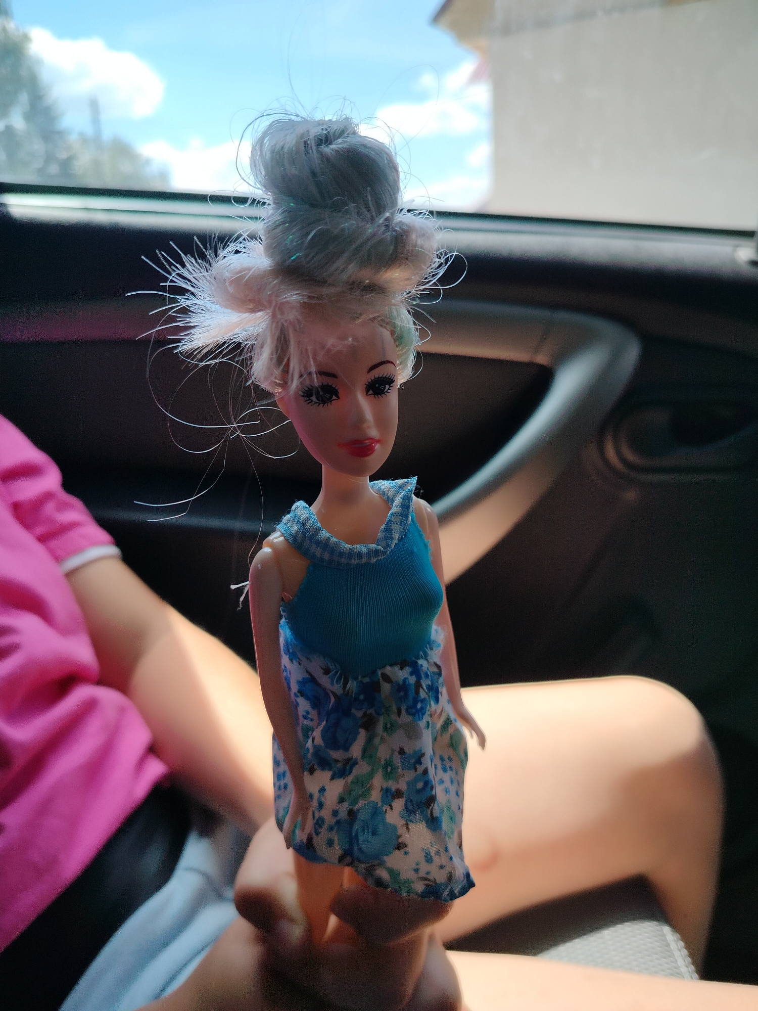 A close-up, eye-level shot shows a doll held in a person's hand, likely inside a vehicle given the visible car interior and window view. The doll, with blonde hair styled in a messy bun and blue floral dress, is the primary subject. Its face features prominent black eyelashes, blue eyes, and red lips. To the left of the doll, a person wearing a bright pink t-shirt is visible, with their arm and leg also in frame. The background consists of a car's interior with a dark handle and dashboard, and beyond that, a bright sky with scattered clouds and distant buildings. The scene is outdoors, possibly in the city of Блохино, Russia, indicated by the presence of a vehicle and a glimpse of an urban or suburban environment.