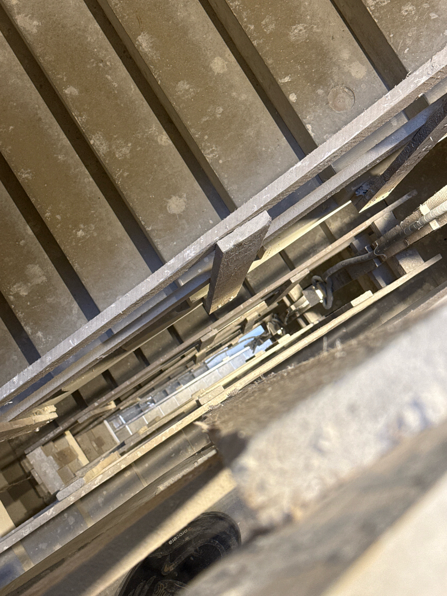 The image depicts a downward view into a concrete stairwell. The stairs are constructed from concrete slabs with metal railings on either side. Visible in the depth of the stairwell are electrical conduits and pipes. At the very bottom of the frame, a portion of a black shoe with "PROCERA" text is visible, suggesting a person is standing at the top of the stairwell. The environment appears to be an interior, possibly in an older building, given the rough concrete texture and visible infrastructure. There is no specific indication of the city of Nowa Sól, Poland within the image itself.