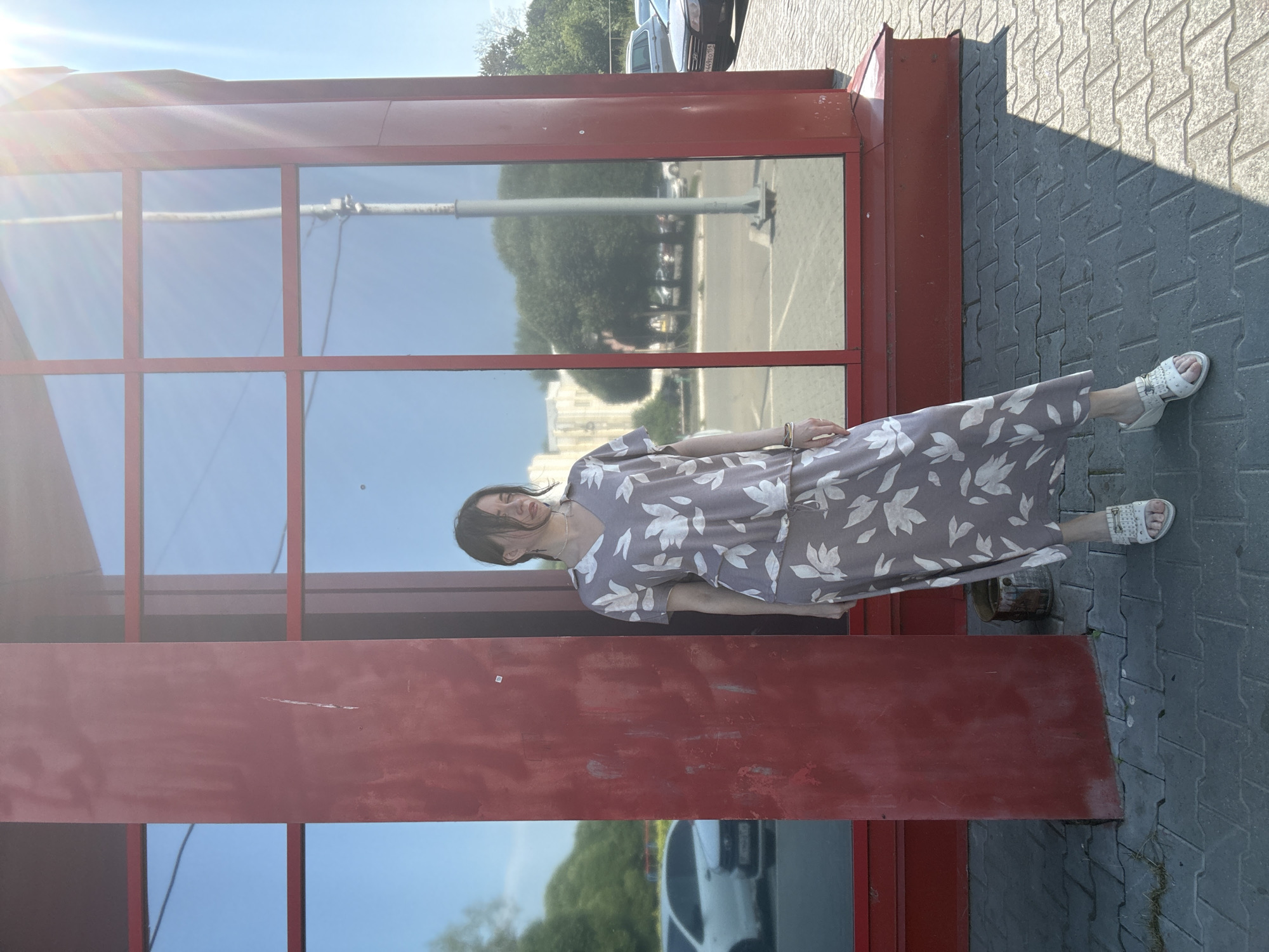 A full-body shot captures a woman standing in front of a red building with large windows. She is wearing a light brown, floral-print dress and white, open-toed sandals. The woman's hair is pulled back, and she is looking down with her eyes closed. The sun is shining brightly, casting a shadow on the ground. The building appears to be located in an urban area, with cars and trees visible in the reflection of the windows. A small, wooden bucket is placed on the ground next to the woman.