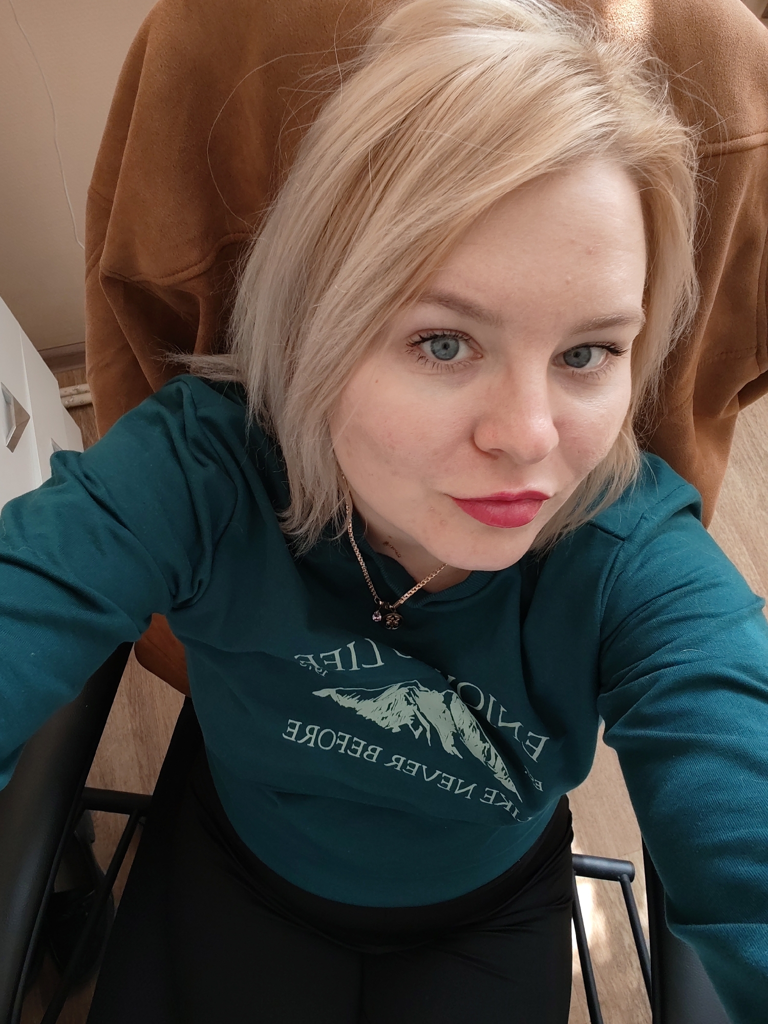 A blonde woman takes a close-up selfie indoors, looking directly at the camera with blue eyes and red lipstick. She appears to be in her 20s or 30s. Her light-colored hair is styled casually. She is wearing a dark teal (or forest green) long-sleeved top with a white graphic and text, and dark, possibly black, leggings or pants. A gold-colored chain necklace with a small pendant is visible around her neck.
The text on her shirt, which is mirrored due to the selfie, reads "ENJOY LIFE LIKE NEVER BEFORE," with a stylized mountain range depicted between "ENJOY LIFE" (above) and "LIKE NEVER BEFORE" (below).
The setting appears to be a room in Izhevsk, Russia, suggested by the provided context. Behind her, a warm brown fabric, possibly part of a chair or jacket, is draped, and a light-colored wall or ceiling is visible in the upper left. Parts of a black chair frame and a wooden floor are visible in the lower half of the image. The lighting suggests it is daytime, with natural light subtly illuminating the scene. Her expression is calm with slightly pursed lips, a common pose for selfies.
