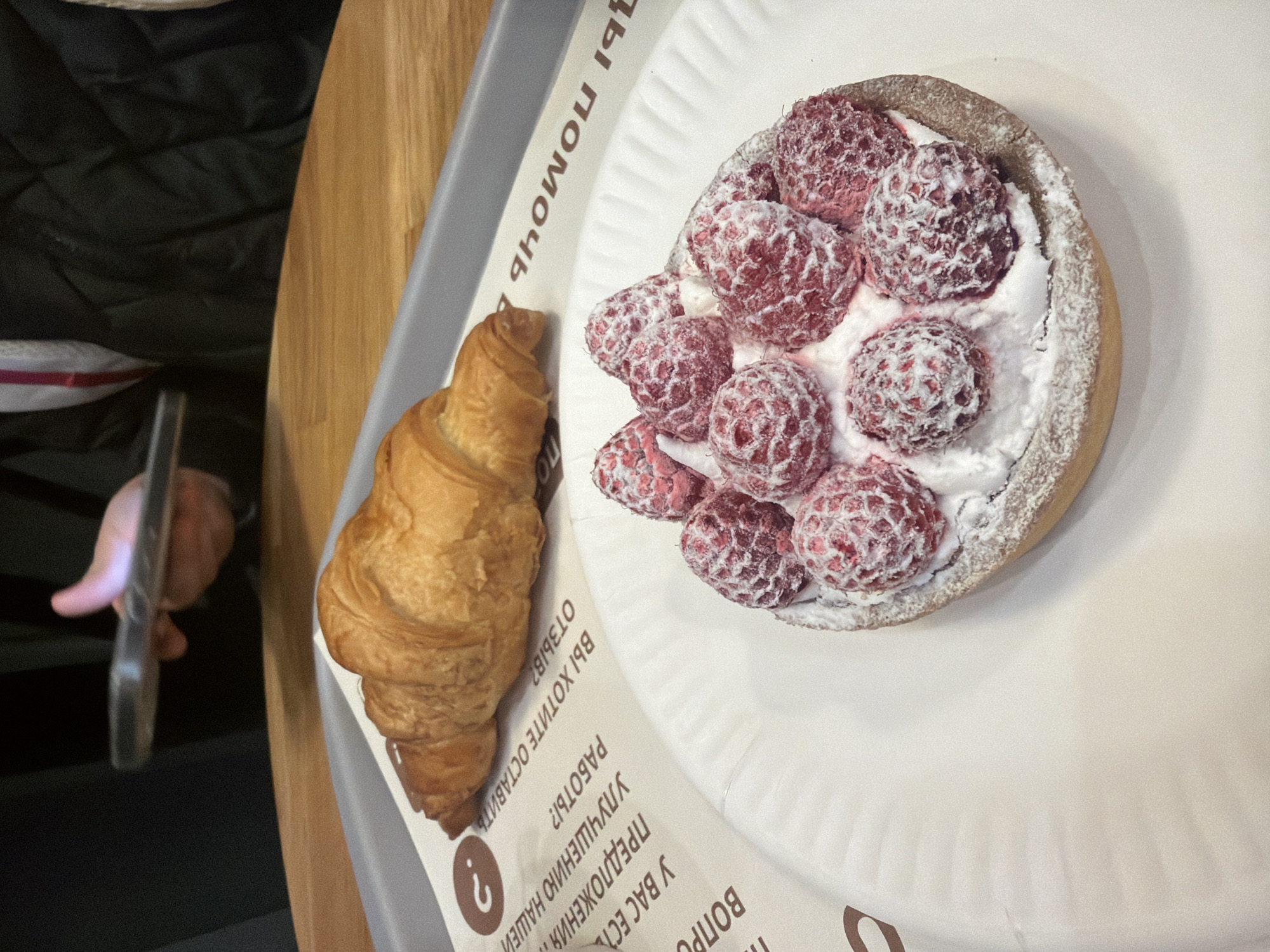The image depicts an indoor scene within a cafe or bakery, likely situated in Saint Petersburg, Russia. In the immediate foreground, a round raspberry tart rests on a white, ridged paper plate. The tart features a pastry crust filled with white cream or mousse, topped with nine whole raspberries heavily dusted with powdered sugar. To the left of the tart and slightly behind it, a golden-brown croissant lies on a grey plastic tray. The tray, along with the tart's plate, is placed on a light-colored wooden table. Beneath the croissant and partially under the tart is a white paper placemat or liner with visible Cyrillic text, including phrases such as "Do you want to leave a review?" and "Suggestions for improving our work?", alongside a brown question mark icon. In the blurred background, a person is visible from the chest up. Their hands hold a dark-colored smartphone horizontally, positioned as if taking a photograph of the food items. The person is wearing a black quilted jacket over what appears to be a garment with white and red stripes. The environment is well-lit, with a focus on the food and the action of photographing it.