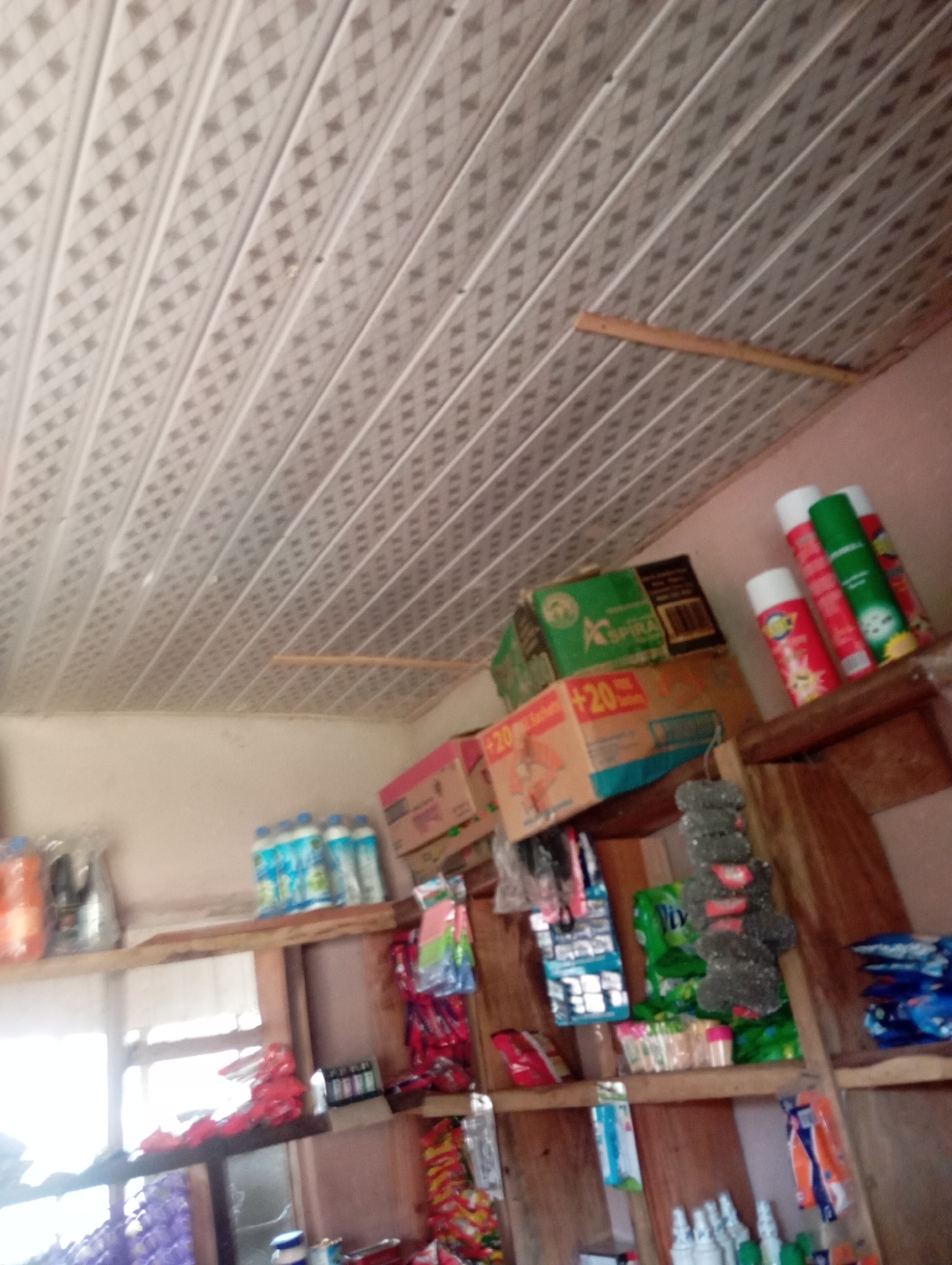 The image depicts the interior of a small convenience store or kiosk, likely located in Jalingo, Nigeria. The scene is crowded with various products displayed on wooden shelves. The ceiling is made of white, patterned panels, and a wooden plank is visible attached to it. On the shelves, there are bottles of what appear to be cleaning supplies or beverages, boxes of packaged goods, aerosol cans, and smaller items packaged in plastic. One shelf holds several bottles of blue liquid, possibly water or detergent. Another shelf has several aerosol cans in red, green, and yellow packaging. Boxes of various sizes and colors are stacked, with some labeled with text and graphics. Various other smaller items in plastic packaging are also visible. The lighting appears to be from an overhead source, possibly fluorescent, casting some shadows and creating a slightly dim atmosphere. The overall impression is of a retail space stocked with a variety of everyday consumer goods. There are no people visible in the image.
